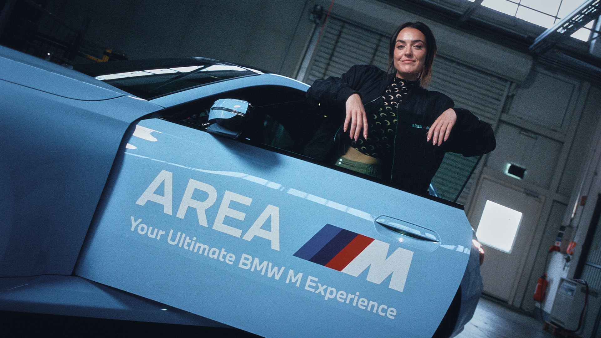 Ein hellblauer BMW Sportwagen mit der Aufschrift 'AREA M Your Ultimate BMW M Experience' auf der Tür. Eine Person in schwarzer Kleidung steht neben dem Fahrzeug in einer Umgebung, die einer Garage oder Werkstatt ähnelt.