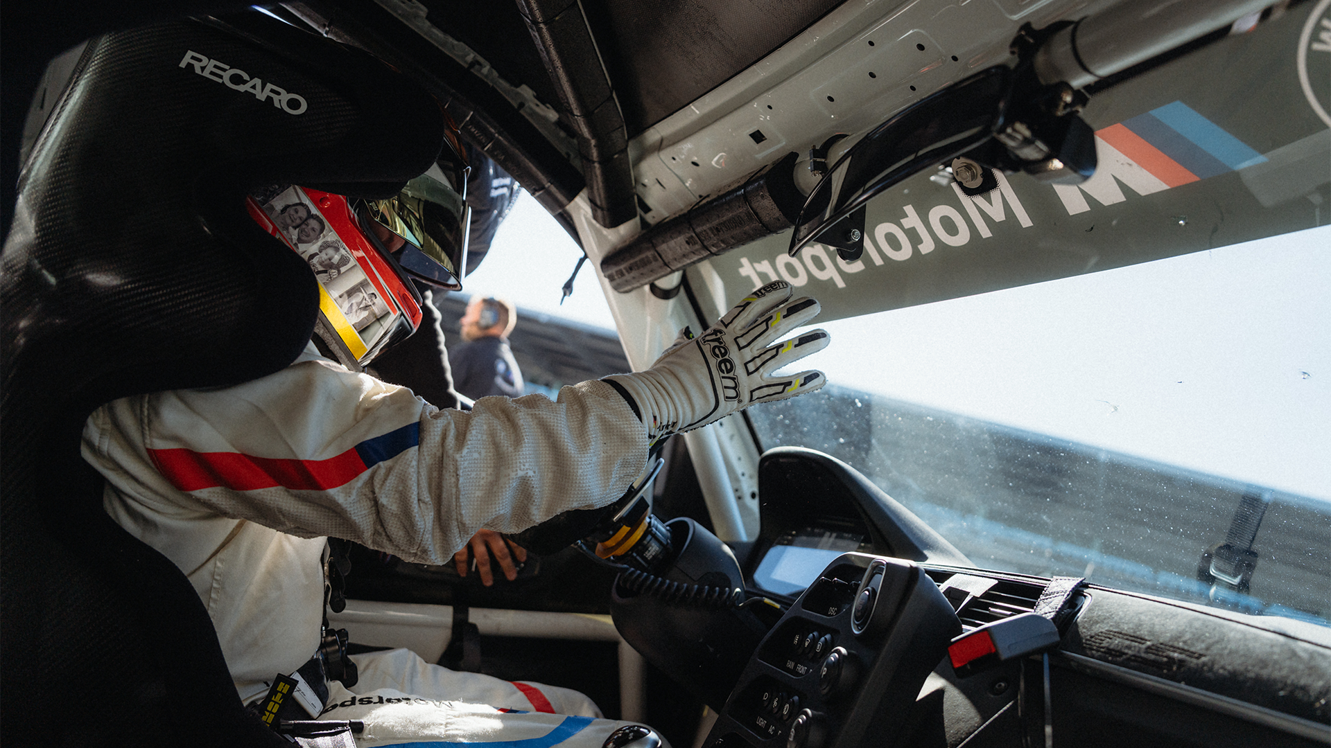 Innenansicht eines Rennwagen-Cockpits mit einem Fahrer, der Rennhandschuhe trägt und in einem Recaro-Sitz sitzt. Das BMW Motorsport Logo ist auf dem Fenster sichtbar, und Rennausrüstung ist im gesamten Innenraum zu sehen.