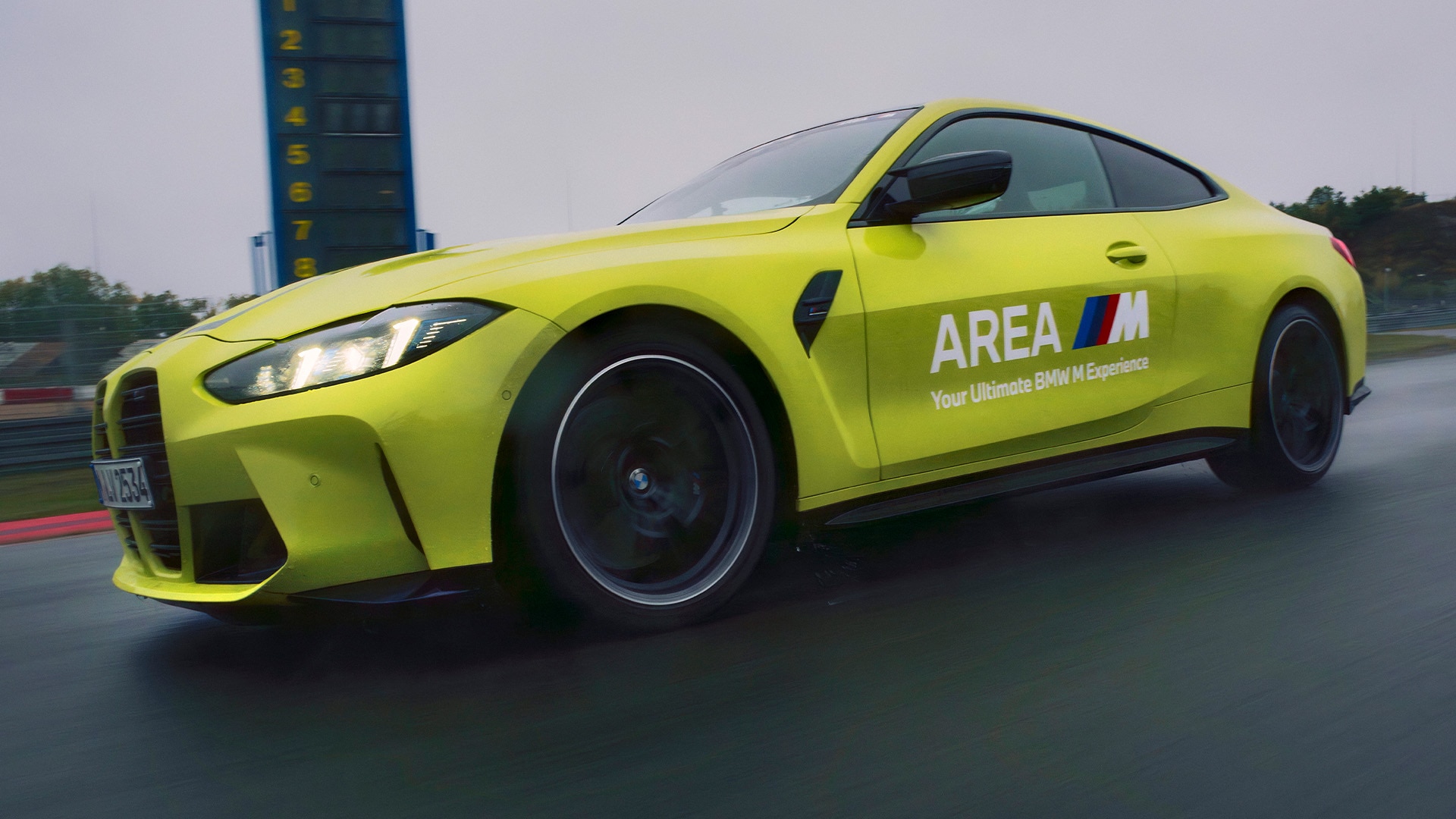Ein leuchtend gelbes BMW M Sportcoupé fährt auf einer Rennstrecke. Das Auto trägt die Aufschrift 'AREA M' und 'Your Ultimate BMW M Experience' an der Seite. Im Hintergrund der Strecke ist ein Zeitturm mit den Nummern 1-8 zu sehen.