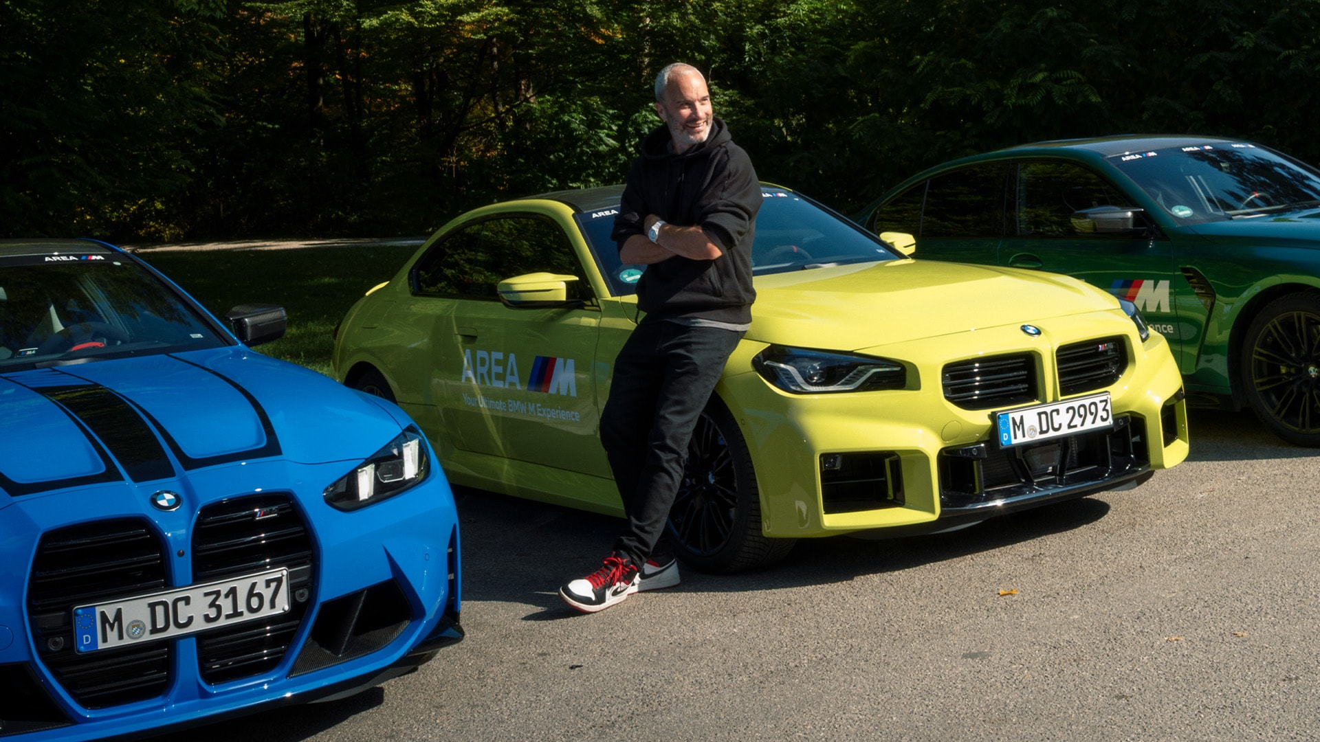 Drei BMW M-Serie Sportwagen in blau, gelb und grün, die auf einer Asphaltfläche vor einem Waldhintergrund parken. Eine Person in schwarzer Kleidung lehnt am gelben Auto, das mit 'AREA M' Branding an der Seite versehen ist.