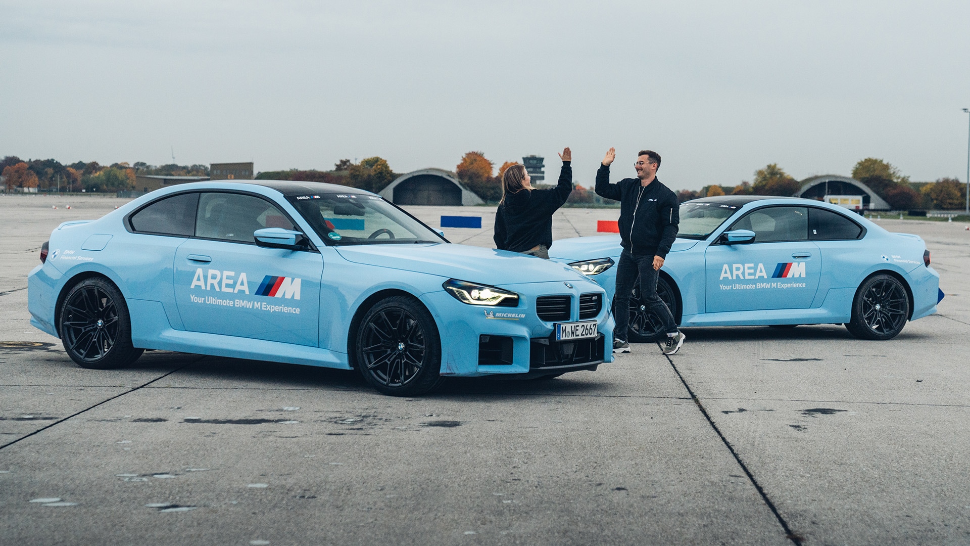 Zwei hellblaue BMW M-Serienfahrzeuge, die auf einem Betonfahrkurs geparkt sind. Die Fahrzeuge tragen die Aufschrift 'AREA M Your Ultimate BMW M Experience' an den Seiten. Zwei Personen stehen zwischen den Fahrzeugen und geben sich ein High-Five. Die Szene spielt auf einer Fahrtstrecke mit herbstlichen Bäumen im Hintergrund.