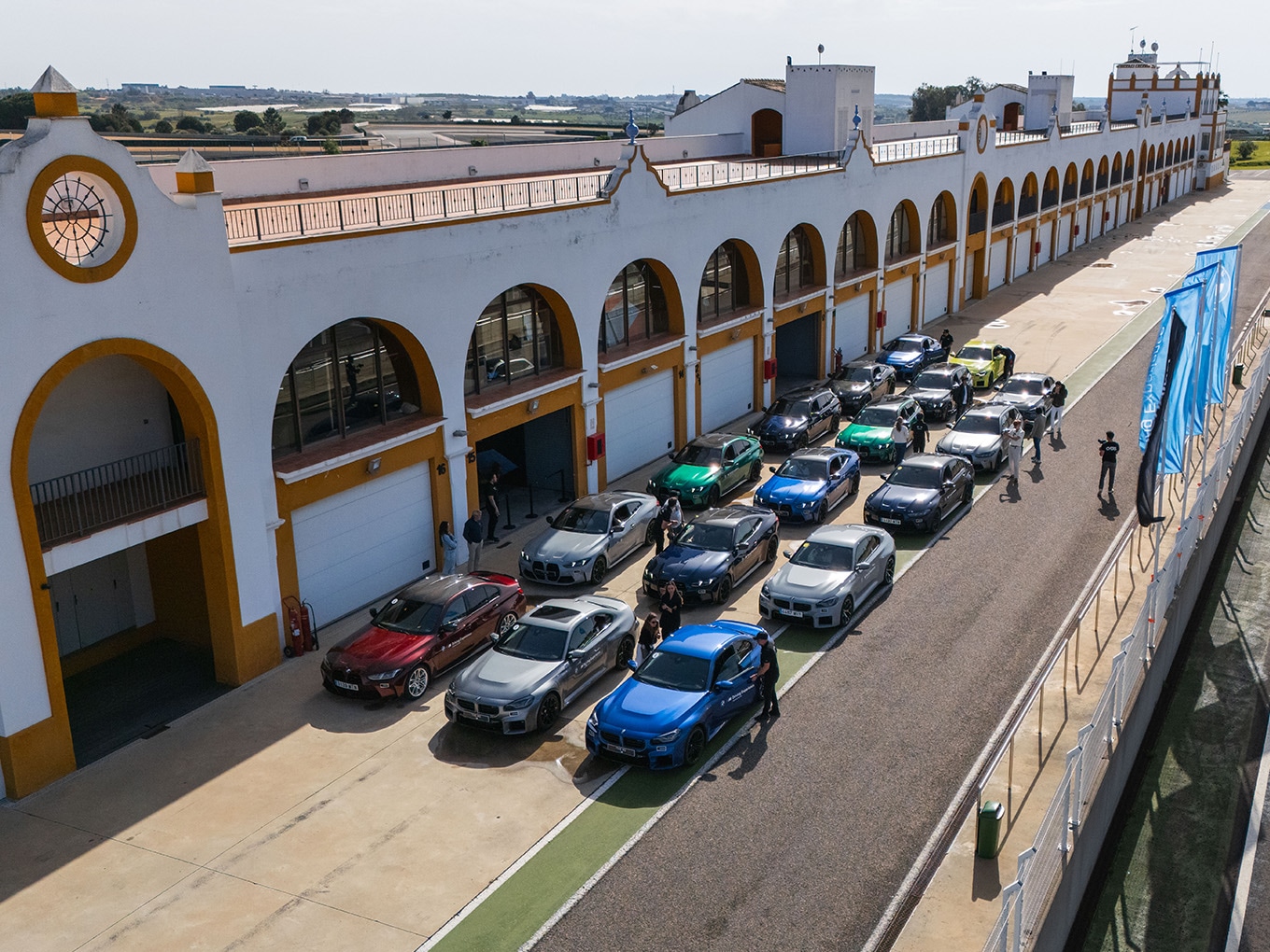 Eine Luftaufnahme eines weißen Gebäudes mit spanischer Architektur, das Torbögen und goldene Verzierungen aufweist. Mehrere BMWs in verschiedenen Farben, sind in Formation auf einer Rennstrecke vor dem Gebäude geparkt. Menschen stehen um die Fahrzeuge herum. Die Anlage scheint Teil einer Rennstrecke zu sein, mit ländlicher Umgebung im Hintergrund.