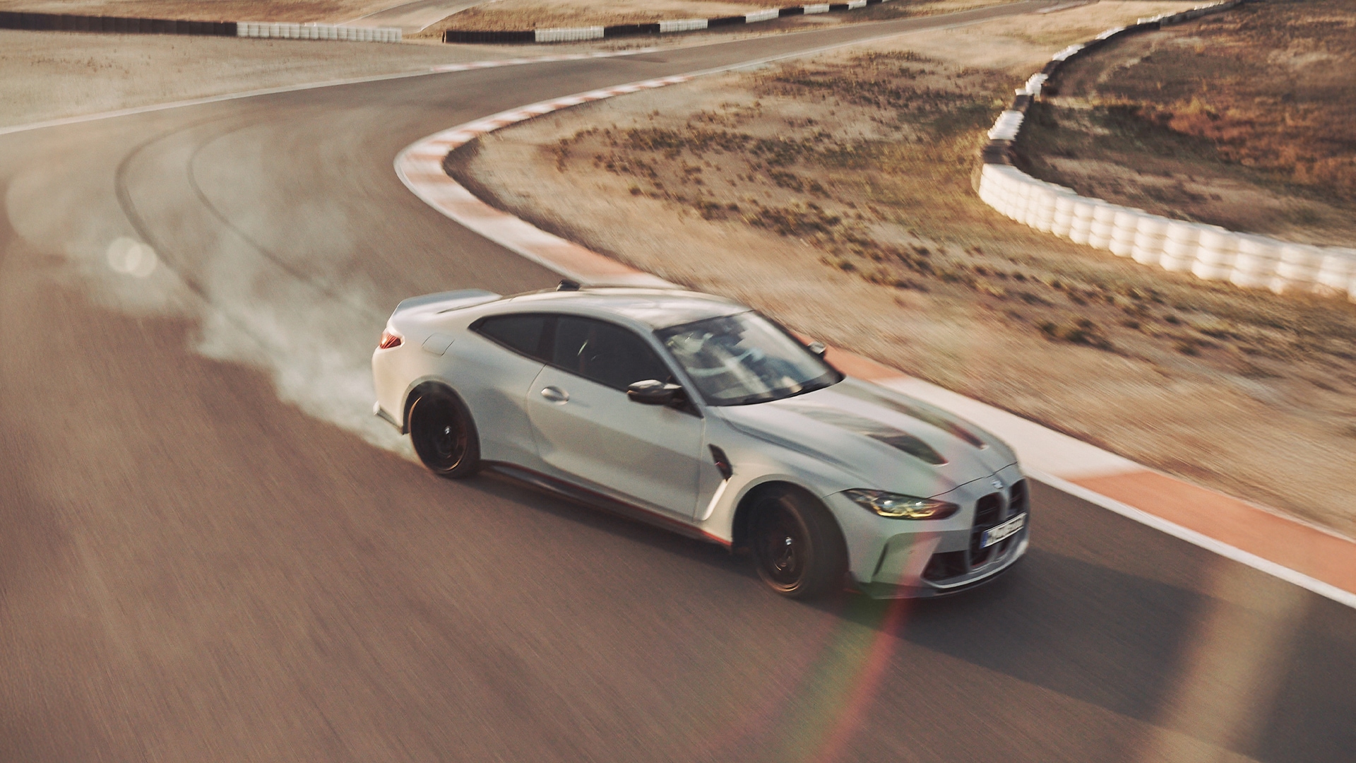 Ein heller BMW Sportwagen driftet um eine Kurve auf einer Rennstrecke und erzeugt dabei Rauch von den Reifen. Das Fahrzeug wird von oben auf einer Asphaltbahn mit wüstenähnlichem Gelände und weißen Sicherheitsbarrieren entlang der Kanten fotografiert.