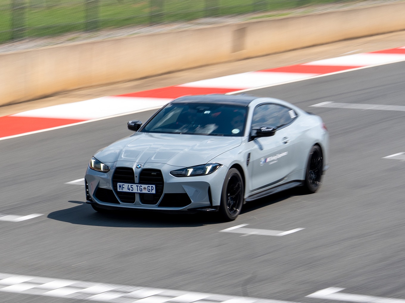 Ein silberner BMW M4 Sportwagen mit schwarzen Felgen fährt auf einer Rennstrecke. Das Auto ist in Bewegung auf einer grauen Asphaltfläche mit rot-weißen Randsteinen am Streckenrand. Das Fahrzeug hat ein südafrikanisches Kennzeichen.