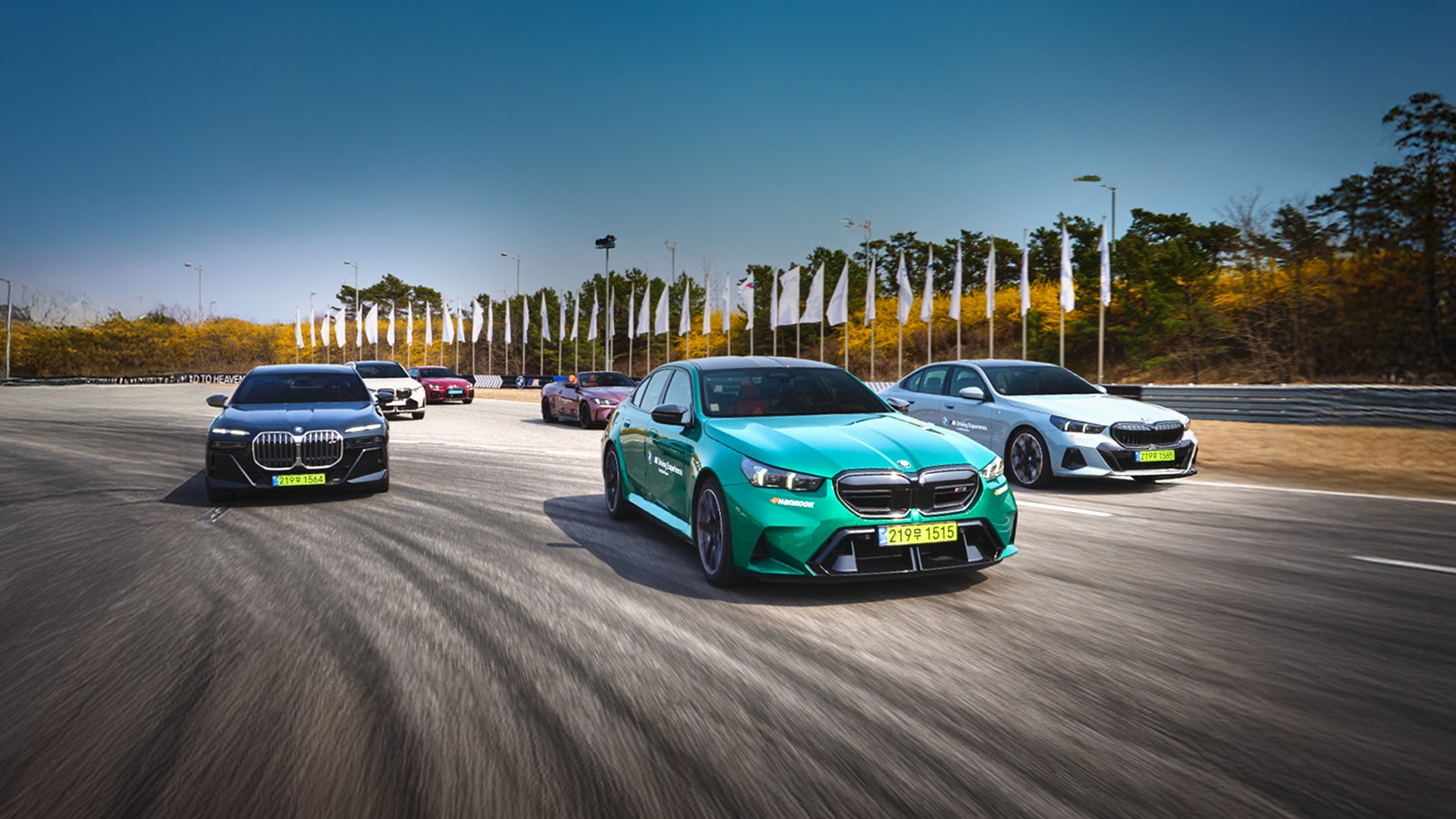 Mehrere BMW Fahrzeuge fahren auf einer Rennstrecke oder einem Testgelände, mit einem türkisfarbenen BMW i4 im Vordergrund. Dahinter befinden sich schwarze, weiße und andere farbige BMW-Modelle. Die Strecke ist mit weißen Fahnen gesäumt und von herbstlichem Laub unter einem klaren blauen Himmel umgeben.