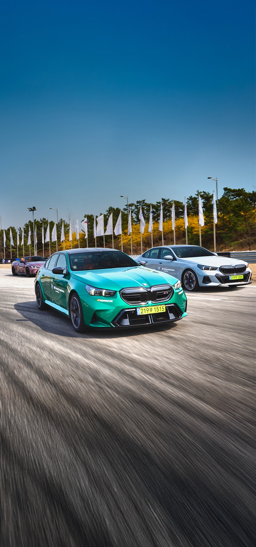 Ein türkisfarbenes Elektrofahrzeug führt ein weißes Auto auf einer Rennstrecke oder einem Testgelände an, mit einem weiteren Auto im Hintergrund. Weiße Fahnen säumen die Strecke vor einem klaren blauen Himmel. Die Bewegungsunschärfe auf der Straßenoberfläche betont die Geschwindigkeit der Fahrzeuge.