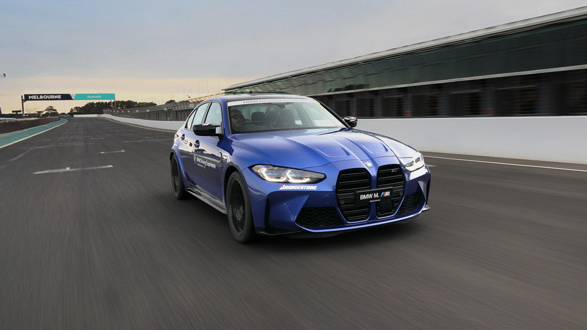 Ein blauer BMW M3 Sportwagen fährt auf einer Rennstrecke. Das Fahrzeug befindet sich auf dem Melbourne-Rundkurs in Australien, wie die Beschilderung zeigt. Das Auto hat die charakteristischen BMW-Nieren und Bridgestone-Sponsoring. Rechts sind die Boxengebäude zu sehen, darüber ein klarer Himmel.
