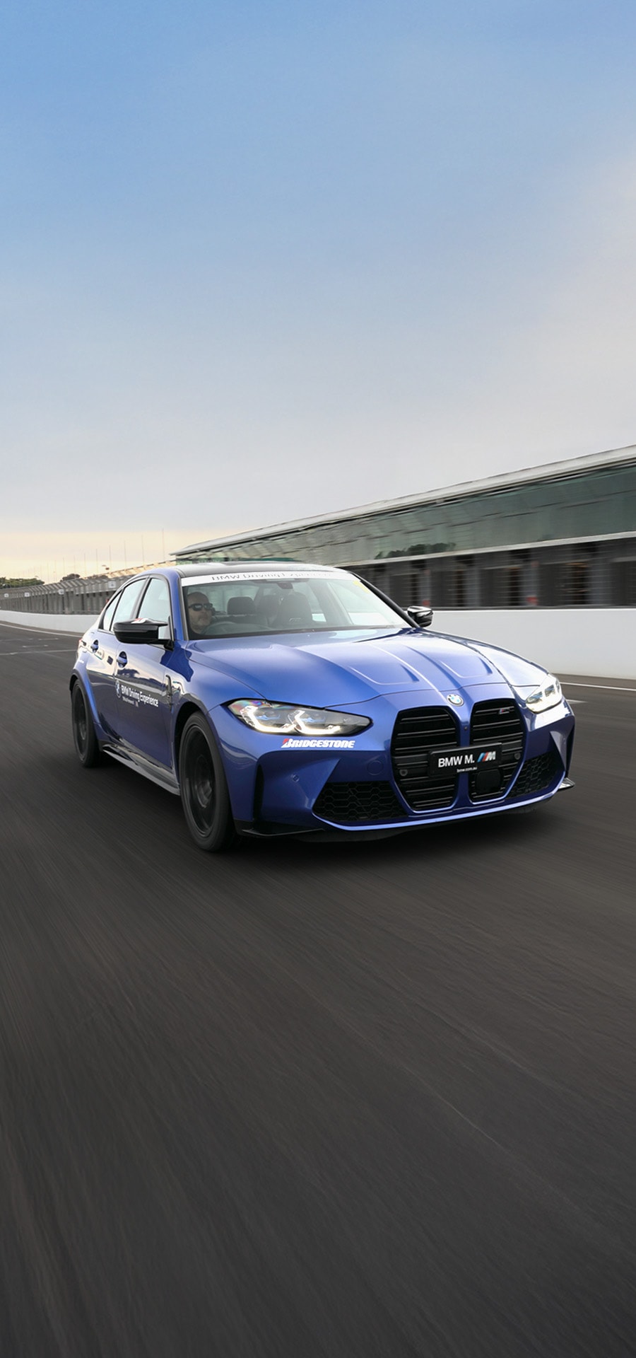 Ein blauer BMW M4 Sportwagen fährt auf einer Rennstrecke mit sichtbaren Boxengebäuden im Hintergrund. Das Fahrzeug verfügt über den markanten großen BMW-Nierengrill und trägt Bridgestone-Branding an der Seite. Das Auto ist in Bewegung vor einem klaren blauen Himmel fotografiert.