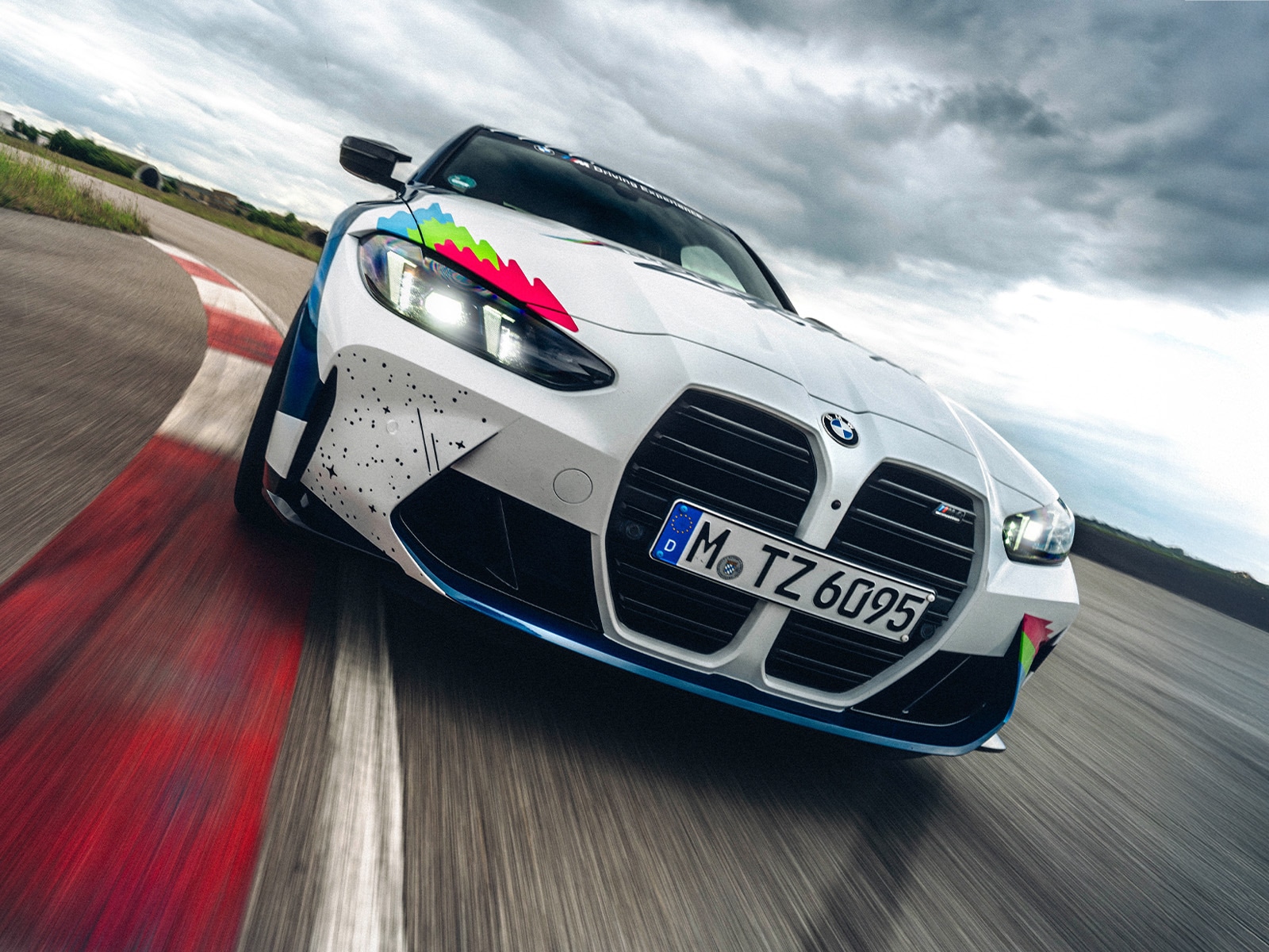 Ein weißer BMW M Sportwagen mit bunten Renngrafikelementen fährt auf einer Rennstrecke. Das Fahrzeug zeigt BMWs markante große Frontgrille und wurde von vorne während der Fahrt auf einer Strecke mit rot-grauen Randsteinen fotografiert. Das Auto hat ein deutsches Kennzeichen und fährt unter einem bewölkten Himmel.