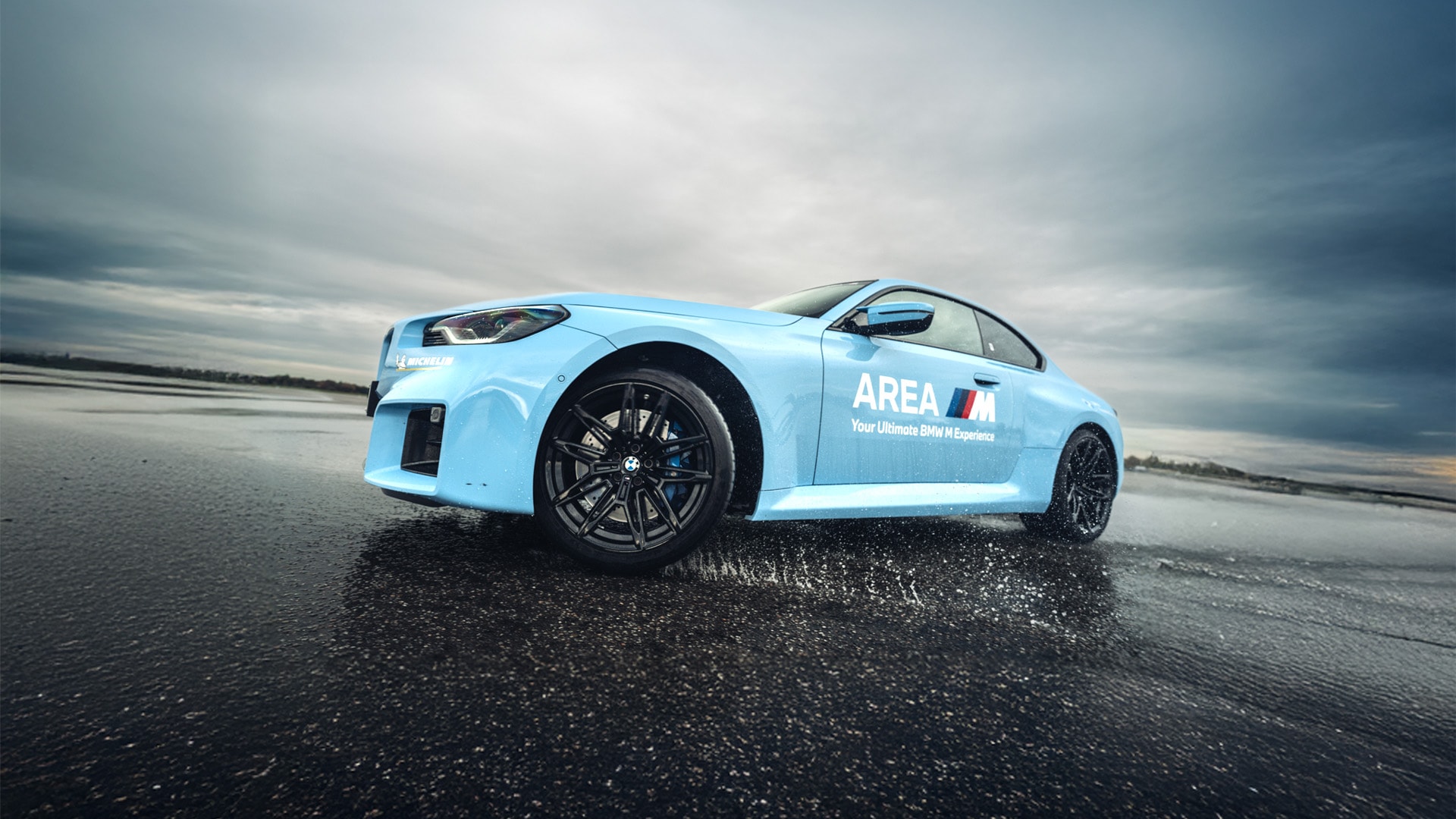 Ein hellblauer BMW Sportwagen mit schwarzen Rädern, der auf einer nassen Asphaltfläche steht. Das Fahrzeug trägt die Aufschrift 'AREA' mit dem Text 'Your Ultimate BMW Experience' an der Seite. Die Szene spielt vor einem dramatischen, bewölkten Himmel, der eine stimmungsvolle Atmosphäre schafft.