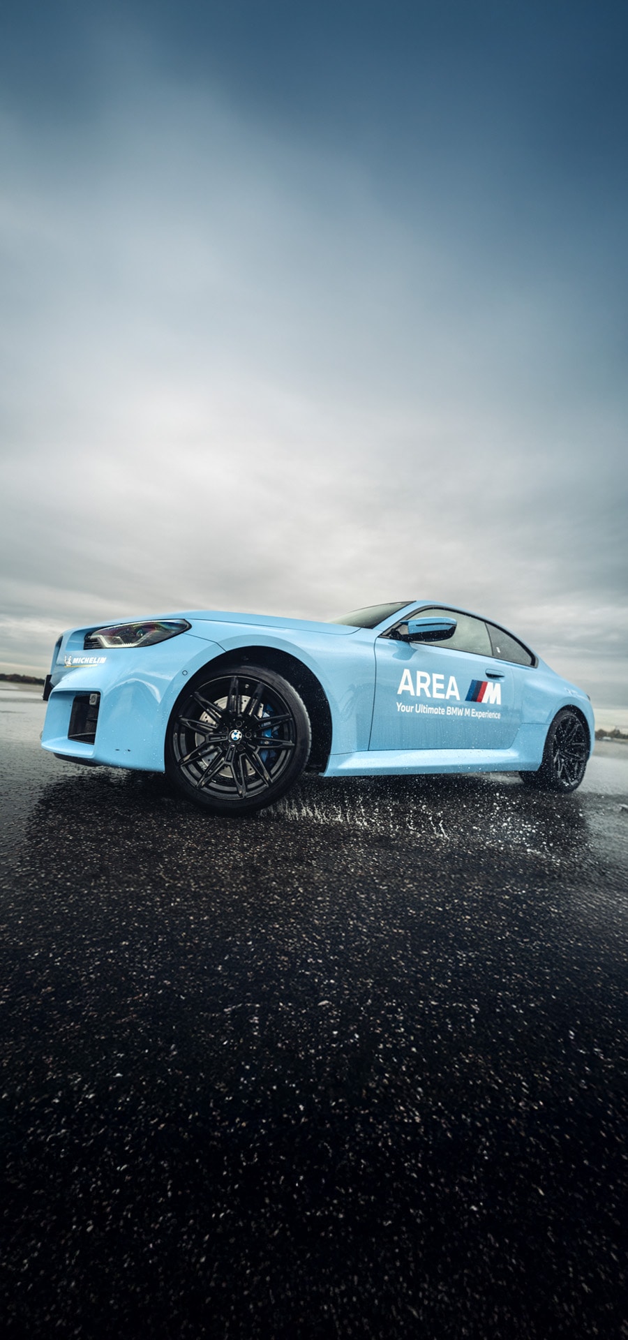 Ein hellblauer BMW Sportwagen mit 'AREA M' Branding an der Seite, der auf einer nassen Asphaltfläche steht. Das Fahrzeug ist mit schwarzen Leichtmetallfelgen ausgestattet und aus einer niedrigen Perspektive vor einem bewölkten Himmel fotografiert.