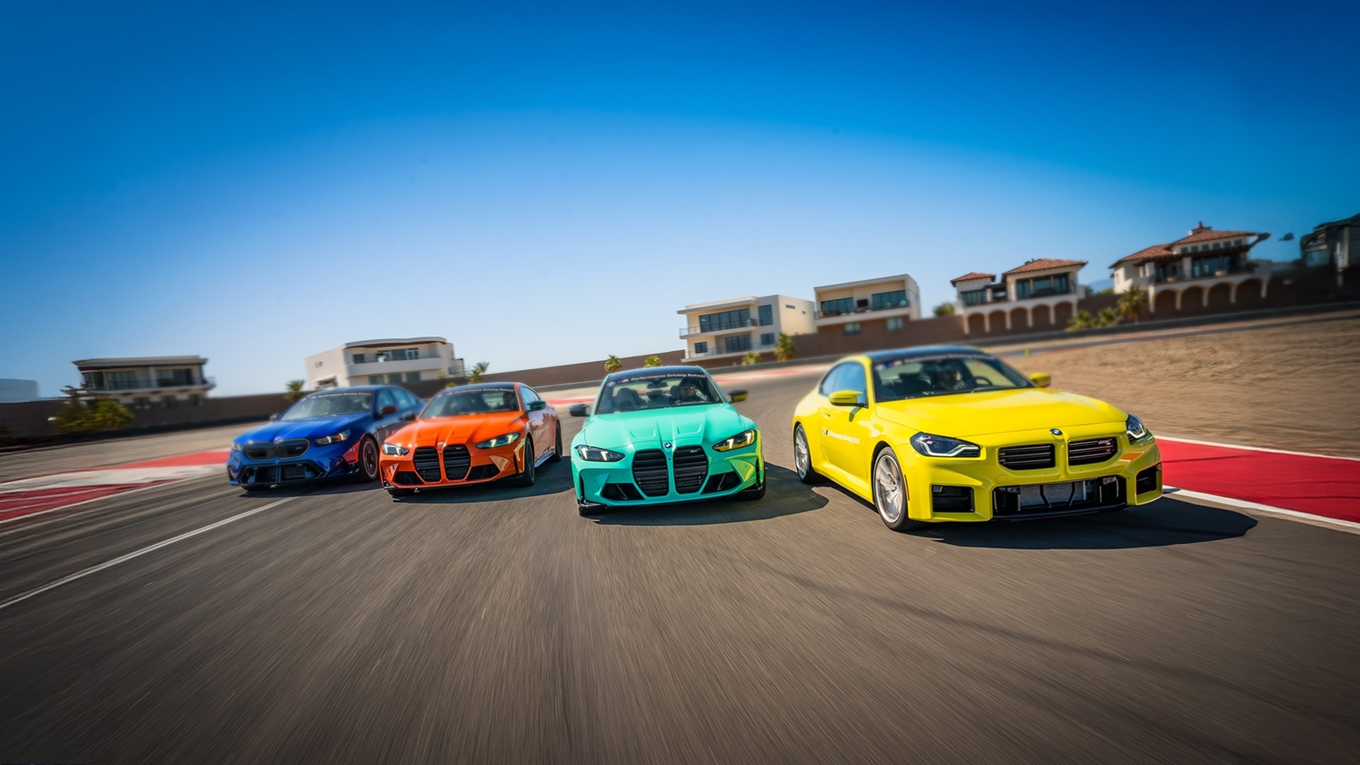 Vier leuchtend farbige BMW Sportwagen fahren in Formation auf einer Rennstrecke. Von links nach rechts: blaue, orange, türkisfarbene und gelbe Fahrzeuge, die auf der Strecke unterwegs sind, mit Luxusgebäuden im Hintergrund unter einem klaren blauen Himmel.