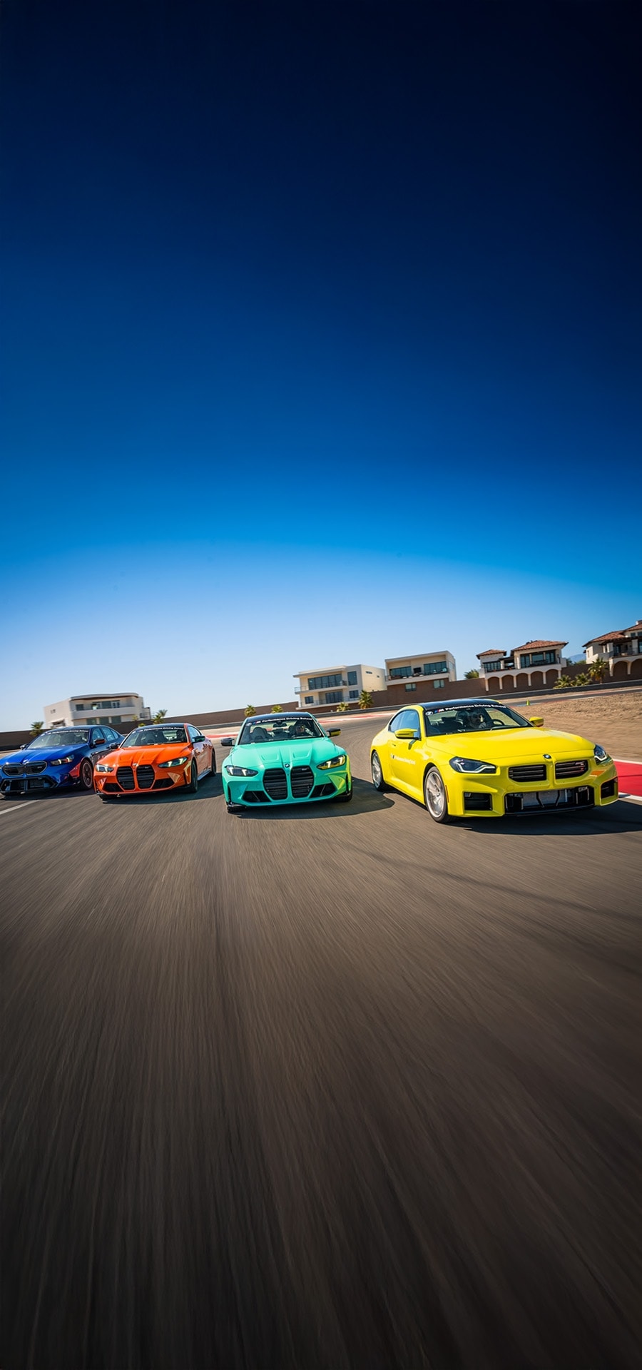 Vier farbenfrohe BMW Sportwagen fahren in Formation auf einer Rennstrecke. Von links nach rechts: blaue, orange, türkise und gelbe Fahrzeuge, die vor einem klaren blauen Himmel mit Gebäuden im Hintergrund fahren.