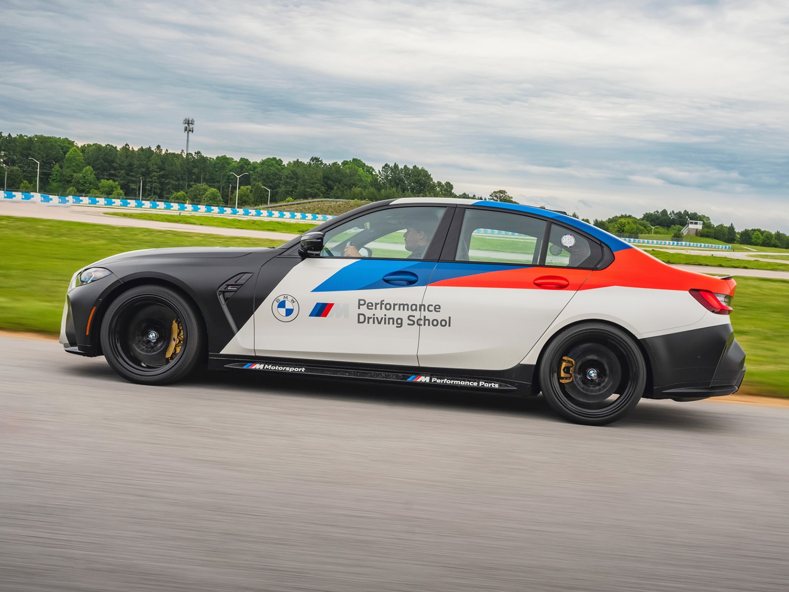 Ein BMW Performance Driving School Fahrzeug in Bewegung auf einer Rennstrecke. Das Auto hat ein markantes Farbschema mit schwarzen Front- und Heckbereichen, weißer Mitte, blauer Fahrerseite und roter Beifahrerseite. Das BMW-Logo und der Text 'Performance Driving School' sind auf der Tür sichtbar. Das Auto fährt auf einer Strecke mit grünem Gras und blauen Barrieren im Hintergrund unter bewölktem Himmel.