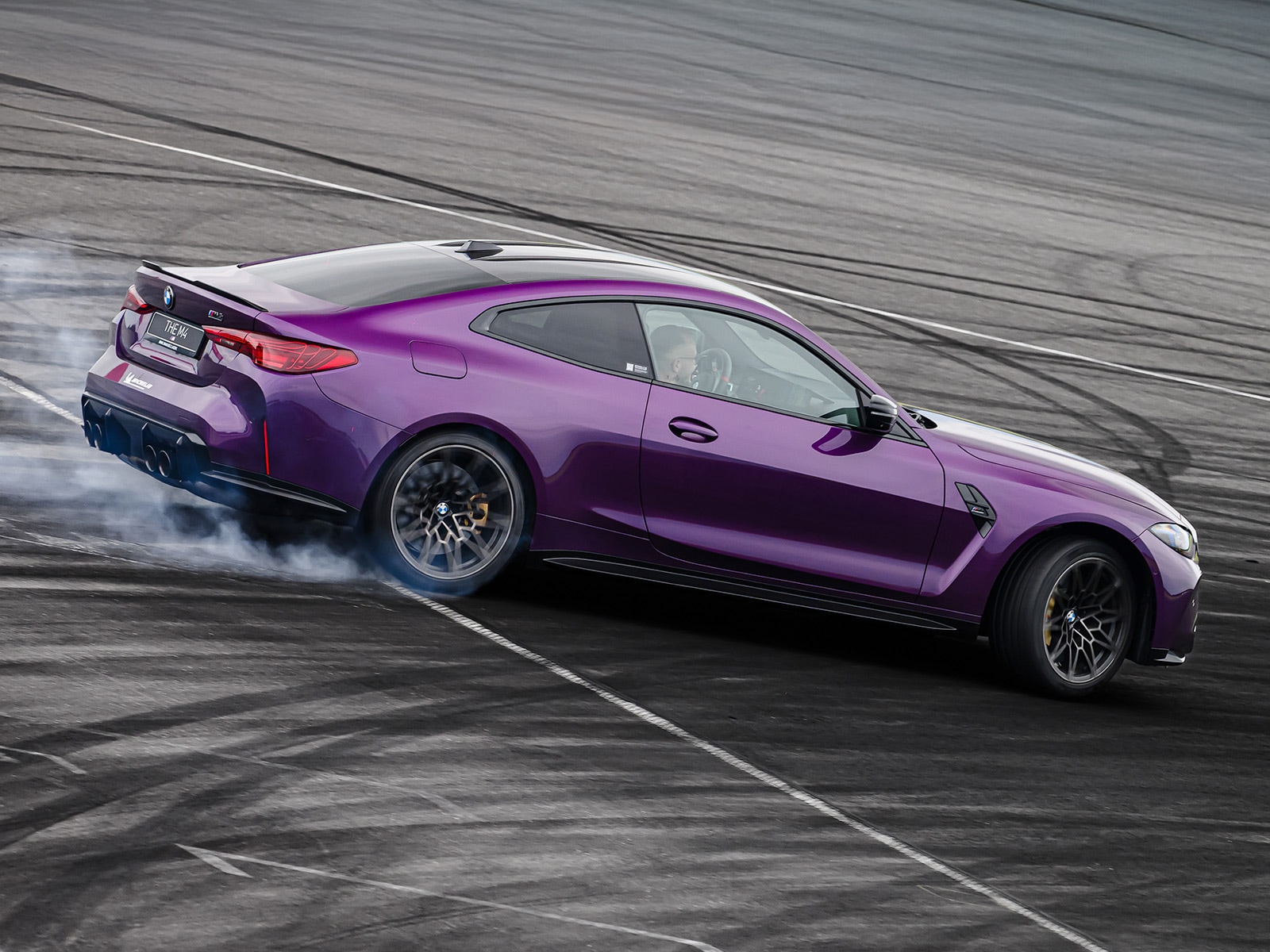 Ein violetter BMW Sportwagen driftet auf einer Rennstrecke und erzeugt Rauch von den Hinterreifen. Das elegante Zweitürer-Coupé hat schwarze Räder und wird von der Seite fotografiert, während es sich auf einer grauen Asphaltfläche mit Reifenspuren bewegt.