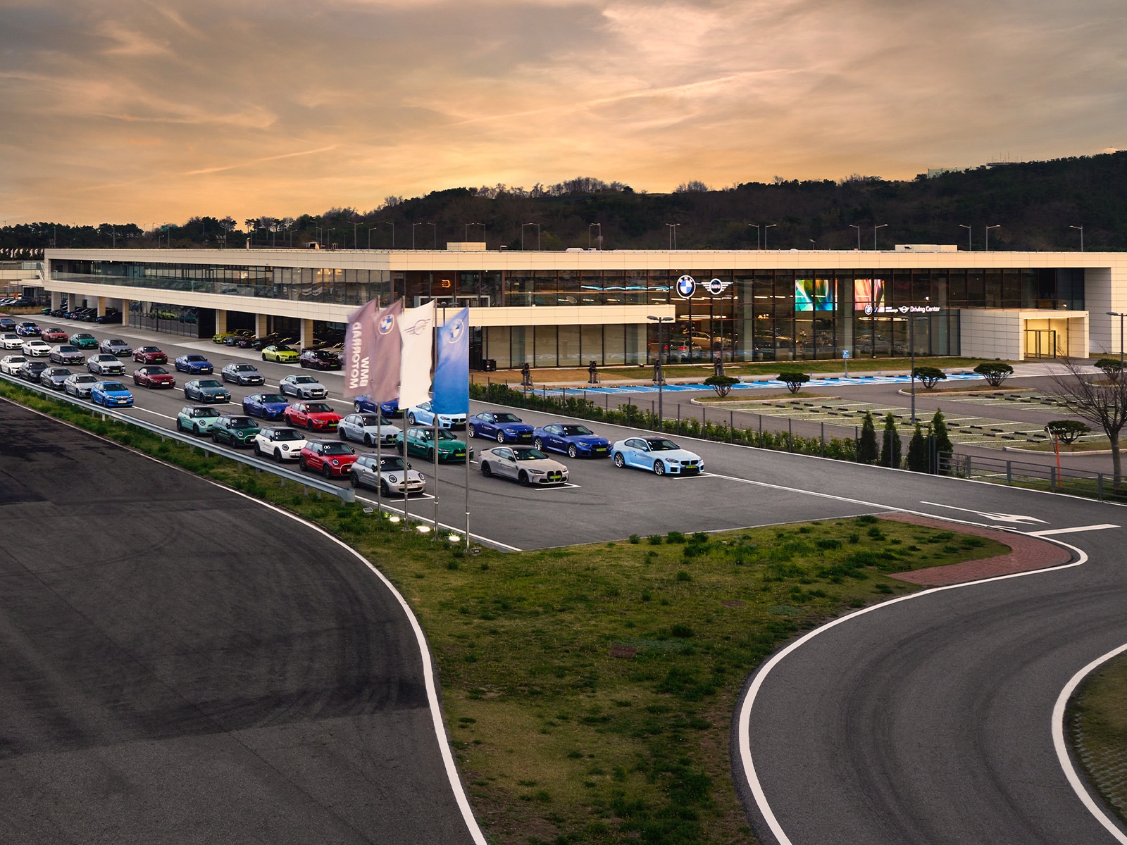 Ein modernes BMW-Autohaus mit einem eleganten Glas- und Betongebäude bei Sonnenuntergang. Im Ausstellungsraum sind die Logos von BMW und MINI zu sehen. Vor dem Gebäude sind zahlreiche farbenfrohe BMW-Fahrzeuge in ordentlichen Reihen geparkt. Das Autohaus befindet sich neben einer Teststrecke oder einem Fahrkurs, mit geschwungenen Asphaltstraßen im Vordergrund. Hügel und Bäume bilden den Hintergrund vor einem goldorangefarbenen Himmel.