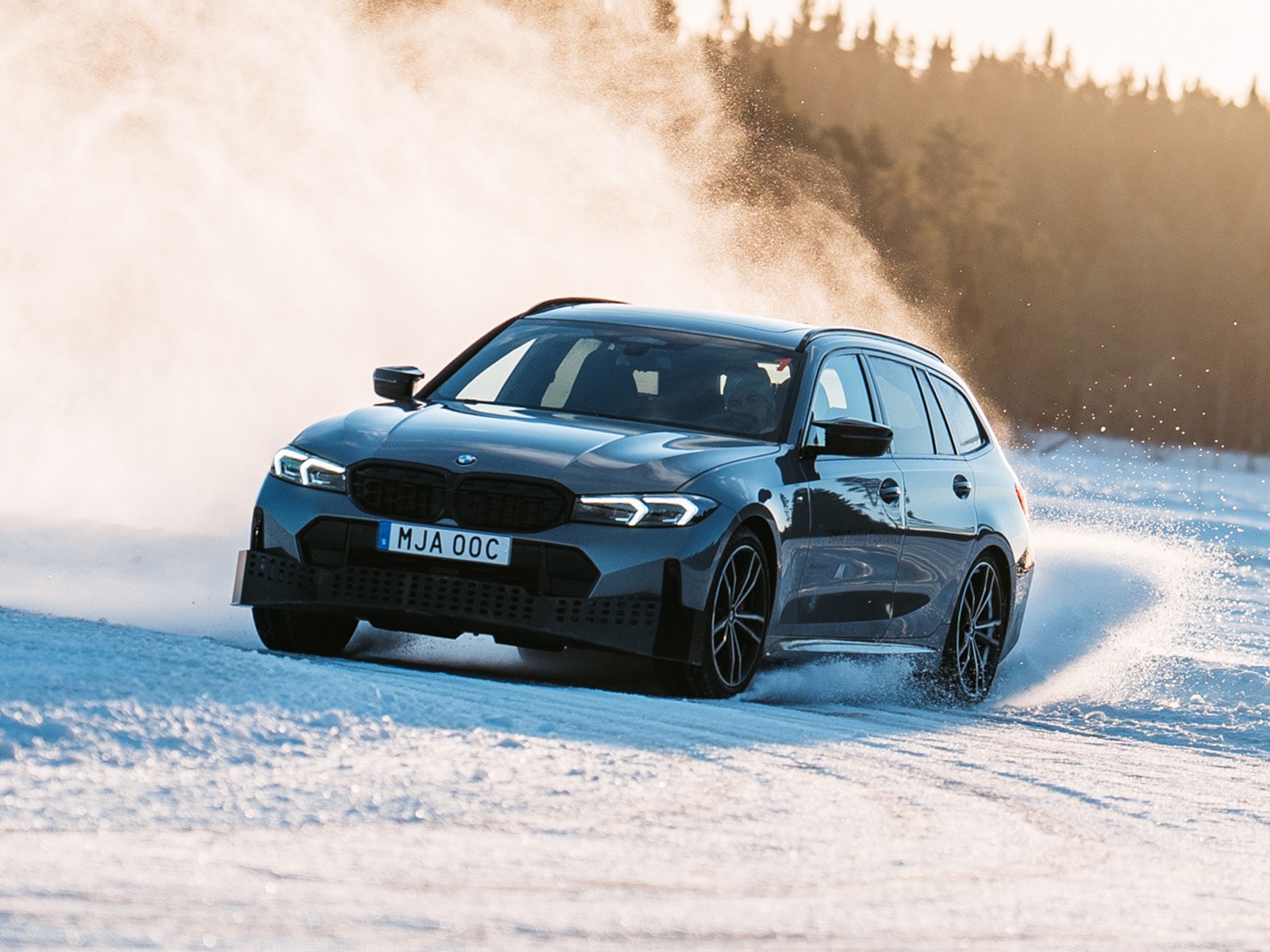 Ein dunkelblauer BMW Kombi fährt durch Schnee und wirbelt Pulverschnee auf, während er sich durch eine Winterlandschaft bewegt. Im nebligen Hintergrund sind Nadelbäume zu sehen.