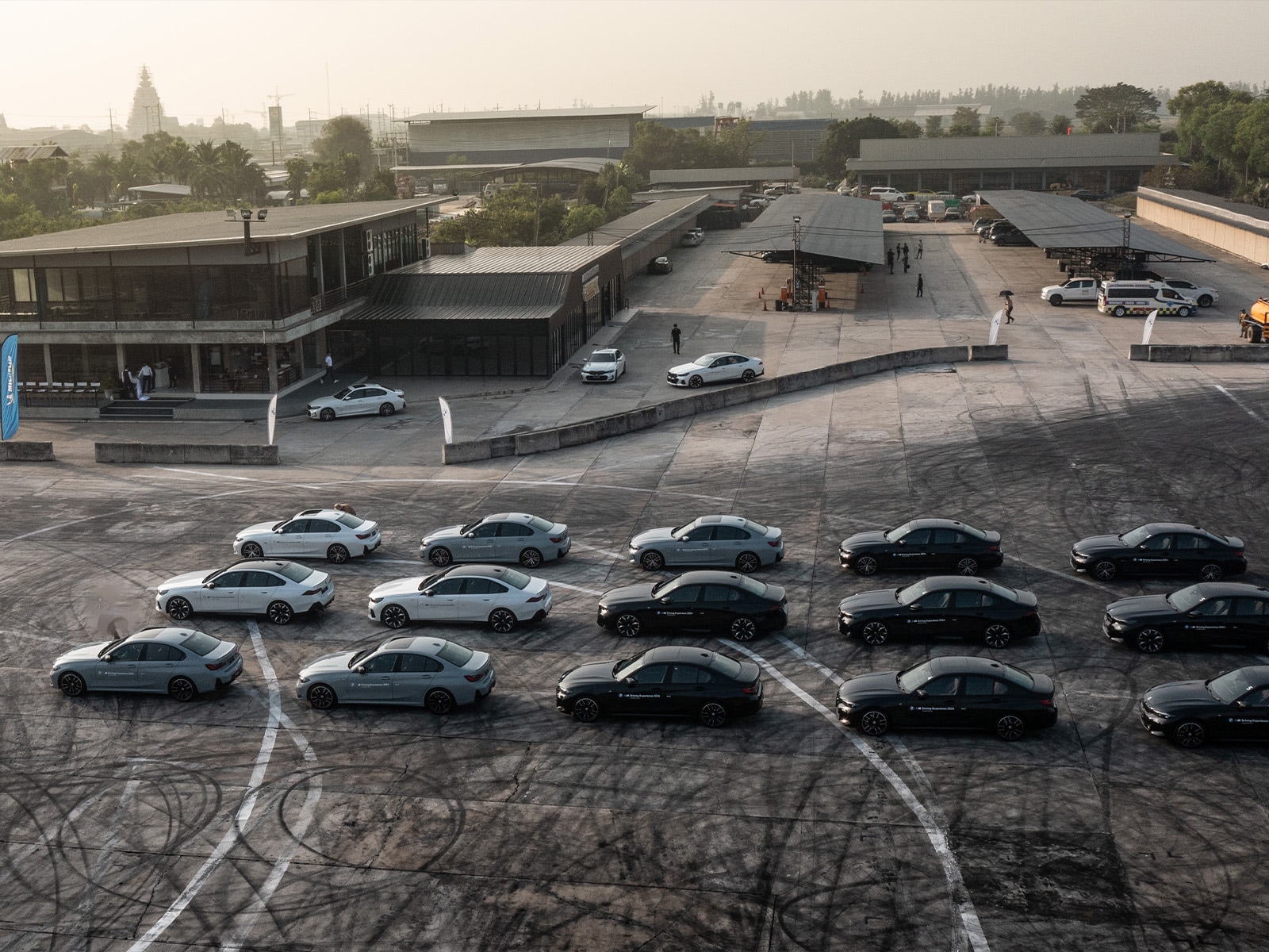 Luftaufnahme eines Gewerbekomplexes mit modernen Gebäuden und einem großen Parkplatz. Mehrere weiße und schwarze Limousinen sind in Reihen auf der Betonfläche angeordnet. Der Komplex umfasst mehrere niedrige Gebäude mit dunklen Dächern. In der Ferne sind Menschen zu sehen, und einige Fahrzeuge parken in der Nähe der Gebäude. Die Szene scheint tagsüber bei dunstiger Atmosphäre aufgenommen zu sein.