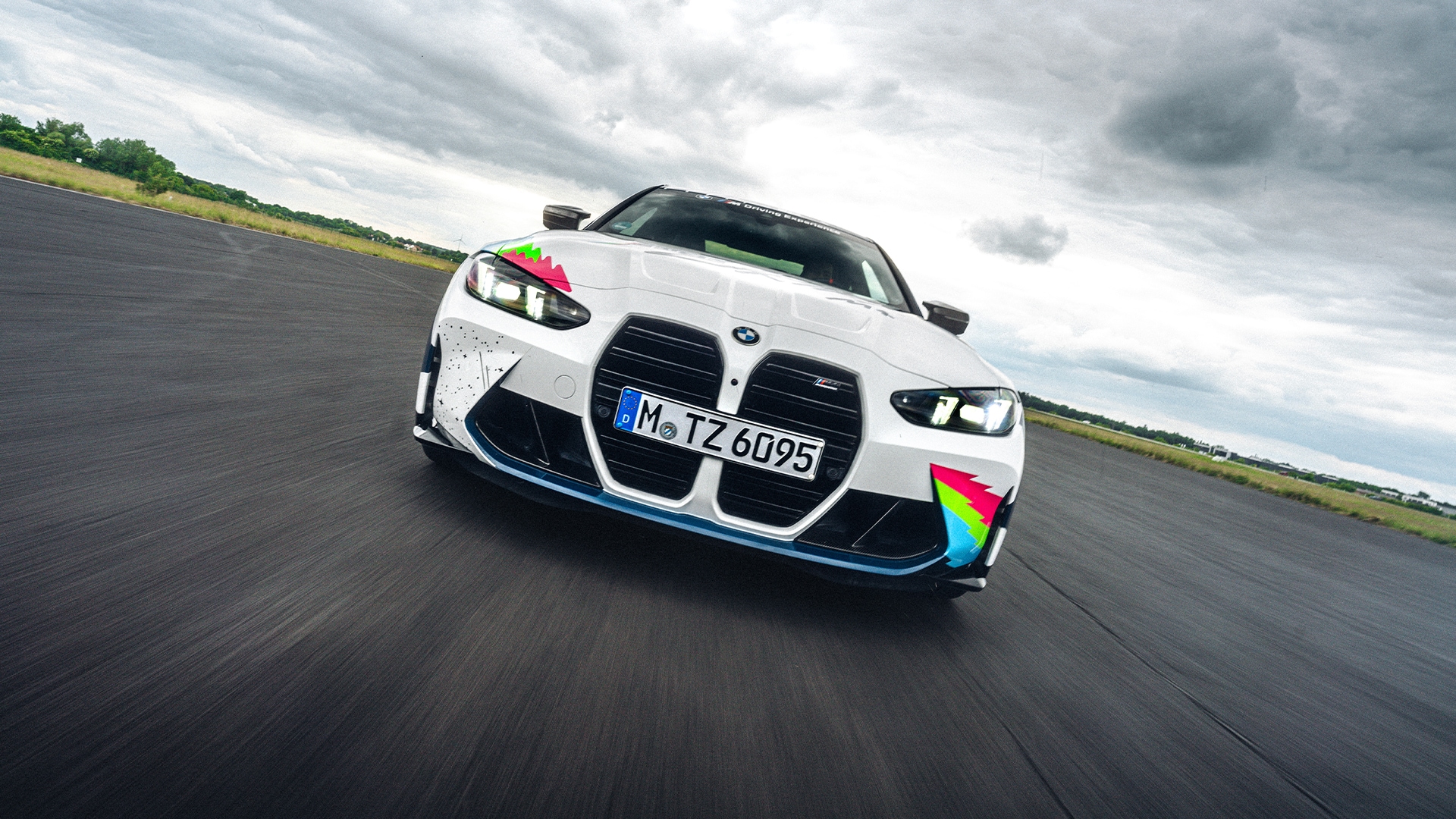 A white BMW sports car with distinctive large kidney grilles and colorful geometric accents on the front bumper, driving on a race track under cloudy skies. The car has a German license plate and is photographed from the front at a dynamic angle that creates a sense of motion.