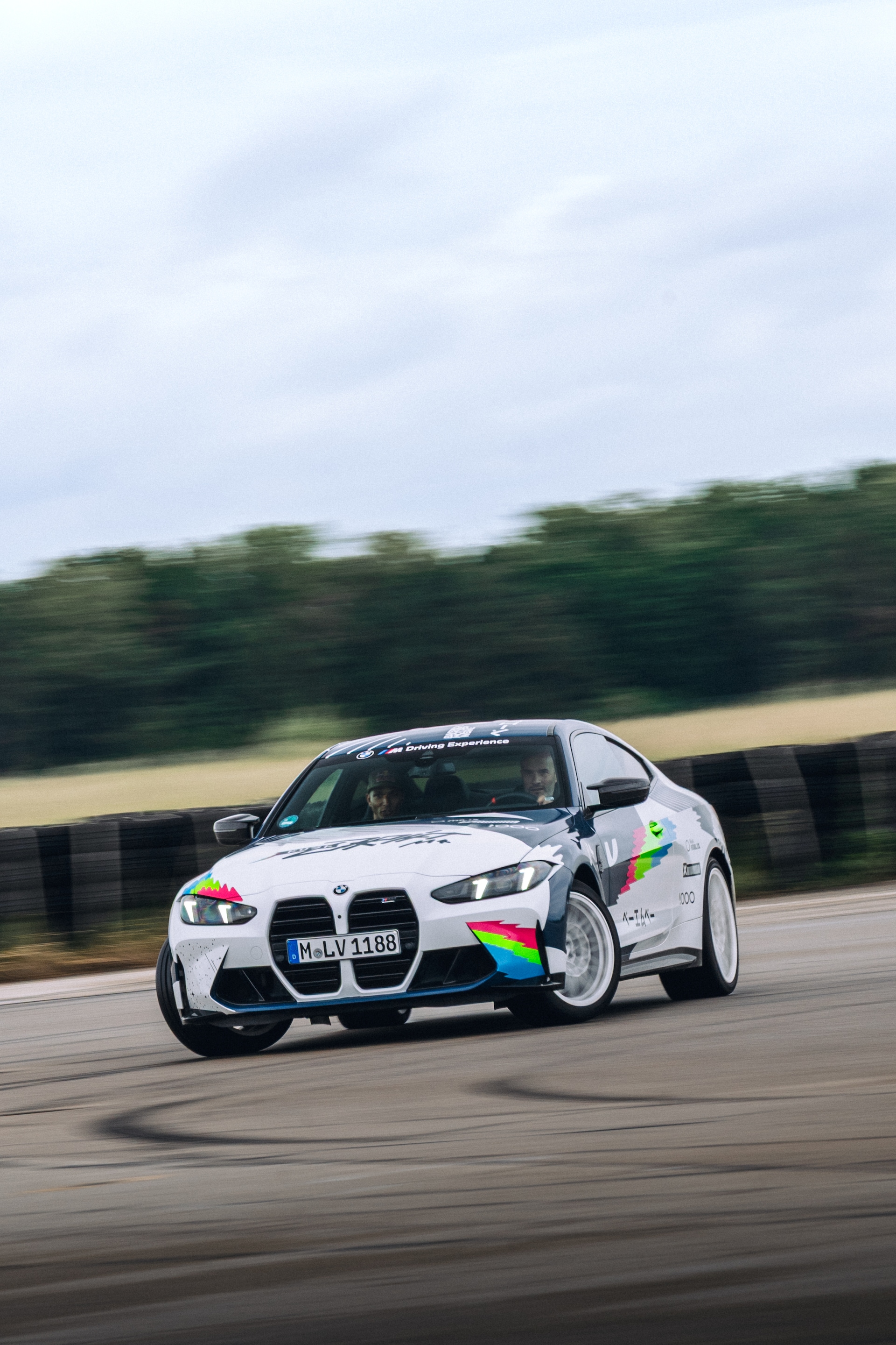 Ein weißer BMW Sportwagen mit bunten grafischen Designs in Bewegung auf einer Rennstrecke. Das Fahrzeug hat das Kennzeichen 'M-LV 1188' und zeigt regenbogenfarbene Akzente an der Seite. Das Auto fährt auf einer asphaltierten Strecke mit Bäumen und Feldern im Hintergrund unter einem bewölkten Himmel.