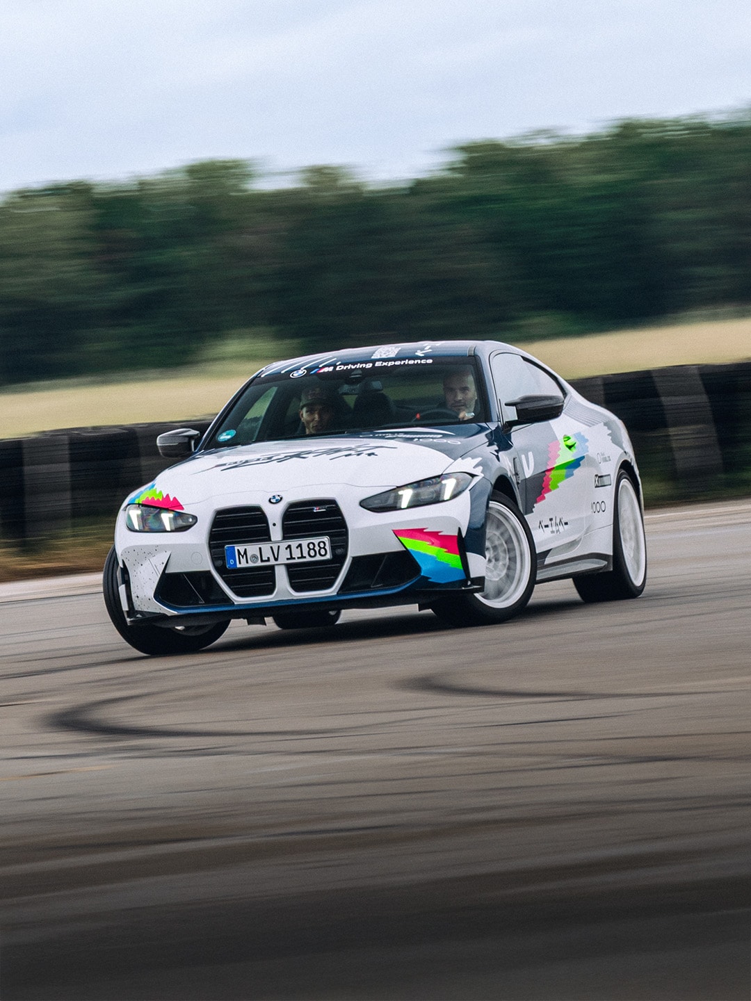 Ein weißer BMW M2 Sportwagen mit bunten Regenbogengrafiken auf der Karosserie, der auf einer Rennstrecke fährt. Das Auto hat ein deutsches Kennzeichen und zeigt Bewegungsunschärfe im Hintergrund mit grünen Bäumen und Feldern.