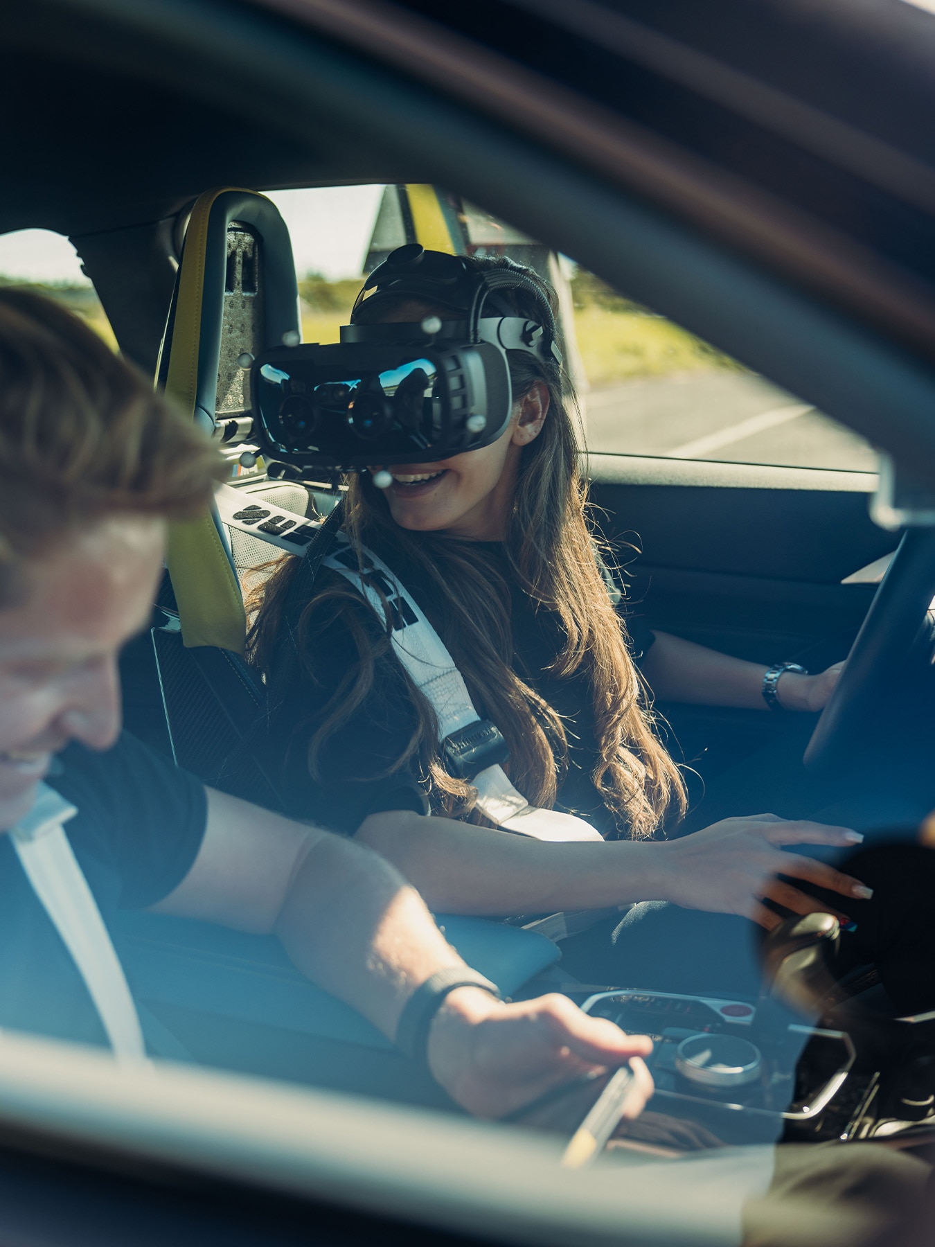 In einem Auto sitzt ein Beifahrer mit einer Virtual-Reality-Brille auf dem Vordersitz, während eine andere Person fährt. Das Bild ist aus der Perspektive aufgenommen, die in das Fahrzeug hineinschaut, wobei durch die Fenster ländliche Gegend zu sehen ist.