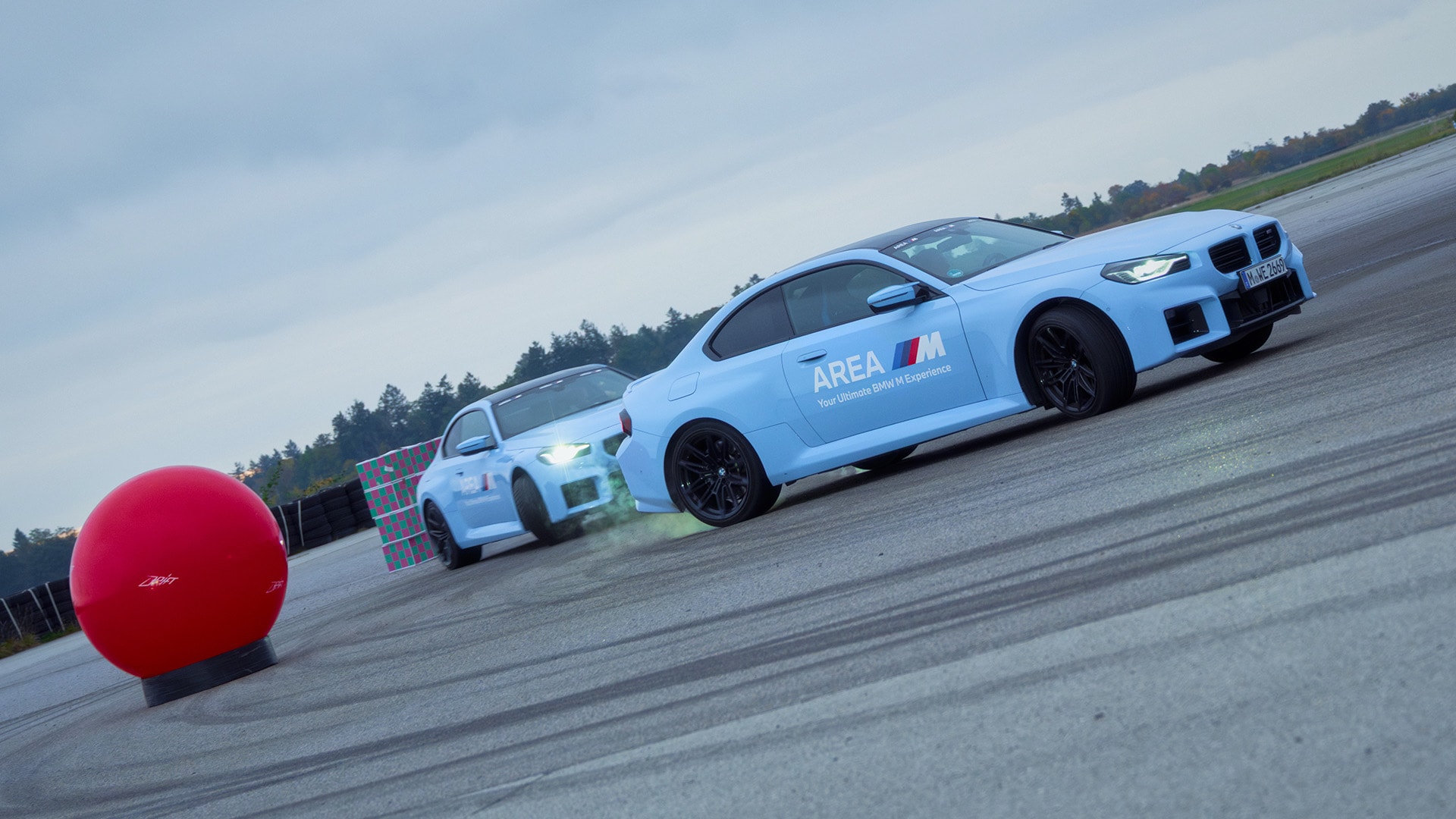 Zwei hellblaue BMW M-Serienfahrzeuge auf einer Fahrstrecke oder Teststrecke. Das vordere Auto zeigt die Aufschrift 'AREA M' auf der Tür. Im Vordergrund ist ein großer roter kugelförmiger Marker zu sehen, mit Reifenspuren auf der Betonoberfläche, die auf Fahrleistungsaktivitäten hindeuten. Im Hintergrund stehen Bäume unter einem bewölkten Himmel.