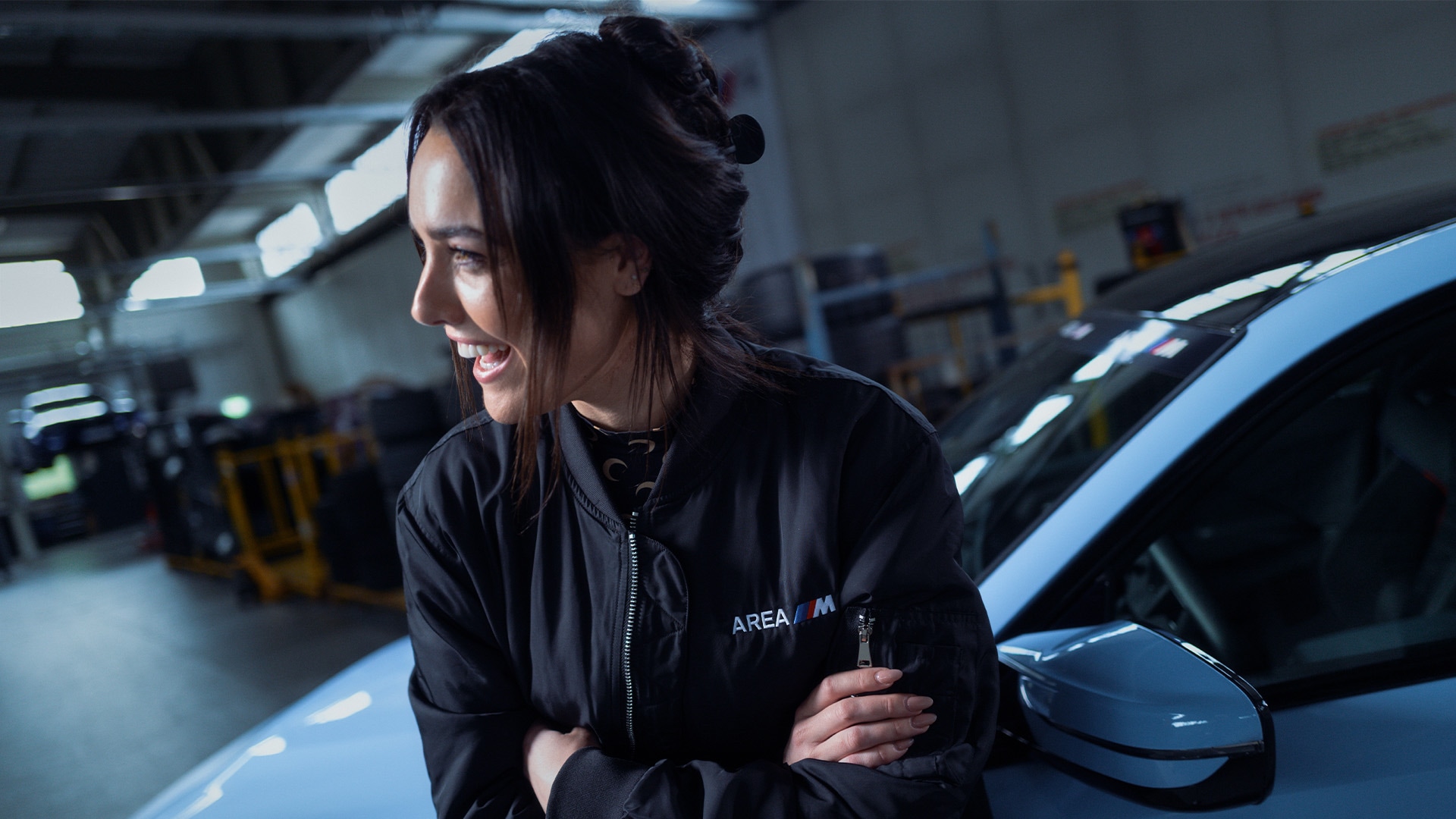Eine Person mit dunklem Haar, das zu einem Dutt frisiert ist, lächelt in einer Autowerkstatt. Sie trägt eine schwarze Jacke mit dem Logo 'AREA M' und steht neben einem blauen Auto. Der industrielle Raum hat Oberlichter und gelbe Sicherheitsbarrieren sind im Hintergrund sichtbar.