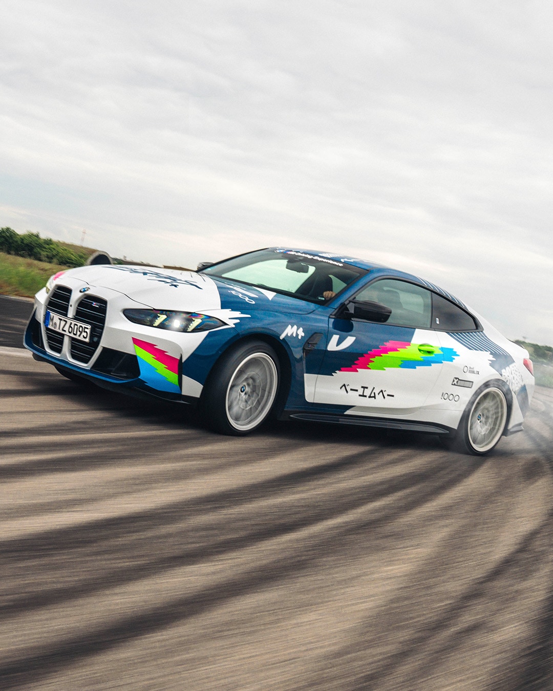 Ein BMW Sportwagen mit buntem Grafik-Design in Blau, Weiß, Pink und Grün driftet auf einer Rennstrecke. Das Auto hat einen markanten Kühlergrill und scheint in Bewegung zu sein, wobei der Boden darunter verschwommen ist. Der Himmel darüber ist bewölkt.
