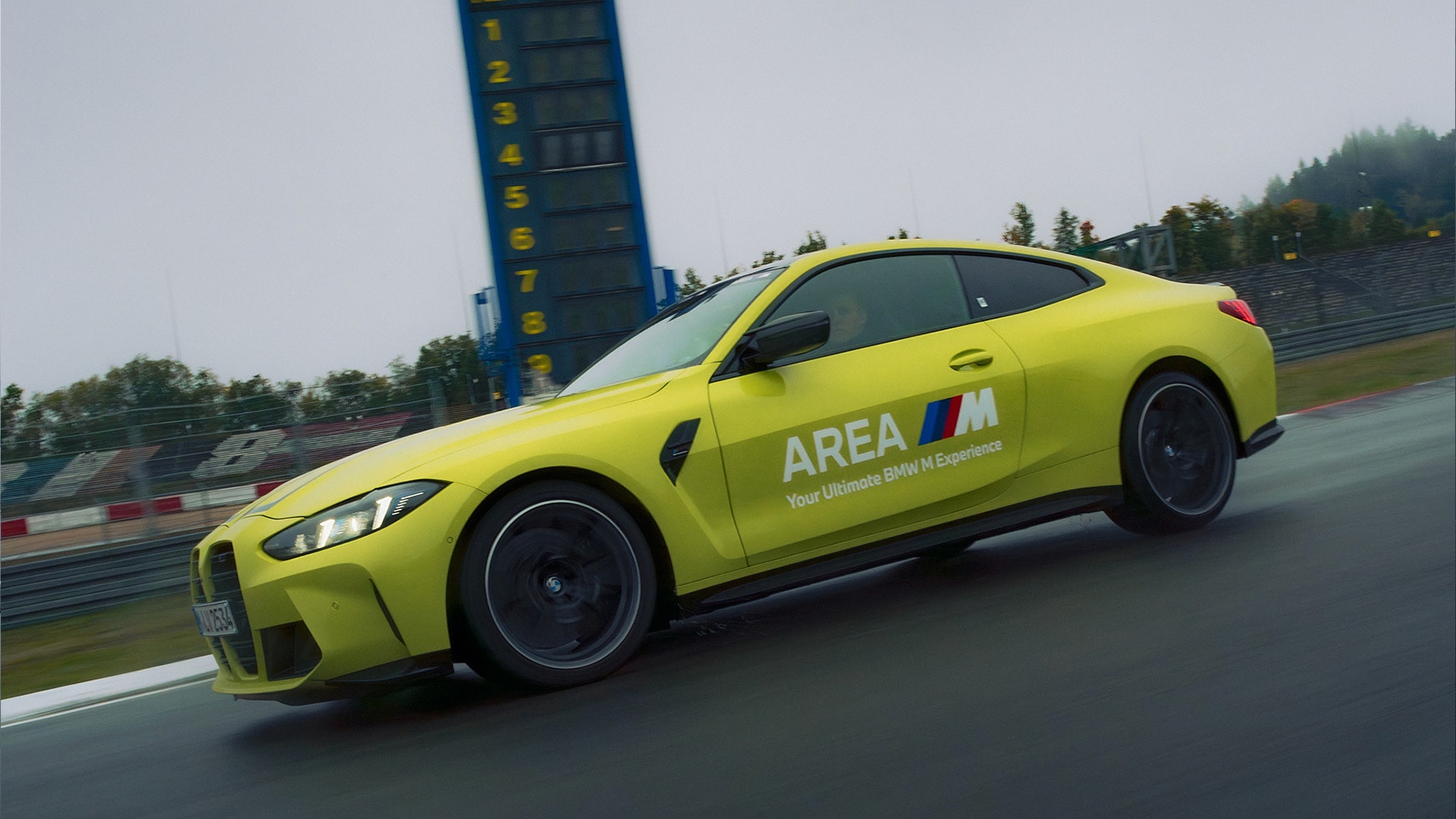 Ein leuchtend gelbes BMW M-Series Sportcoupé fährt auf einer Rennstrecke. Auf der Seite des Fahrzeugs steht 'AREA M Your Ultimate BMW M Experience'. Im Hintergrund sind eine blaue Rundenzähltafel und Rennstreckenbarrieren an einem nebligen Tag zu sehen.