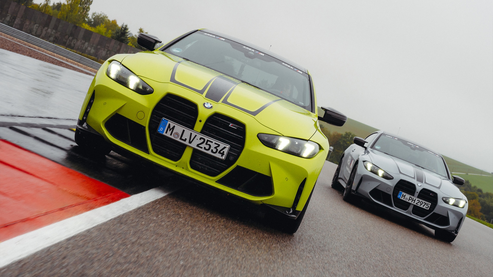 Zwei BMW M3 Sportwagen auf einer nassen Rennstrecke. Das Fahrzeug im Vordergrund ist leuchtend gelbgrün mit Rennstreifen, während ein graues Modell dahinter folgt. Beide Fahrzeuge zeigen BMWs charakteristisches Frontgrill-Design und befinden sich auf einer Strecke mit sichtbarer rot-weißer Bordsteinkante.