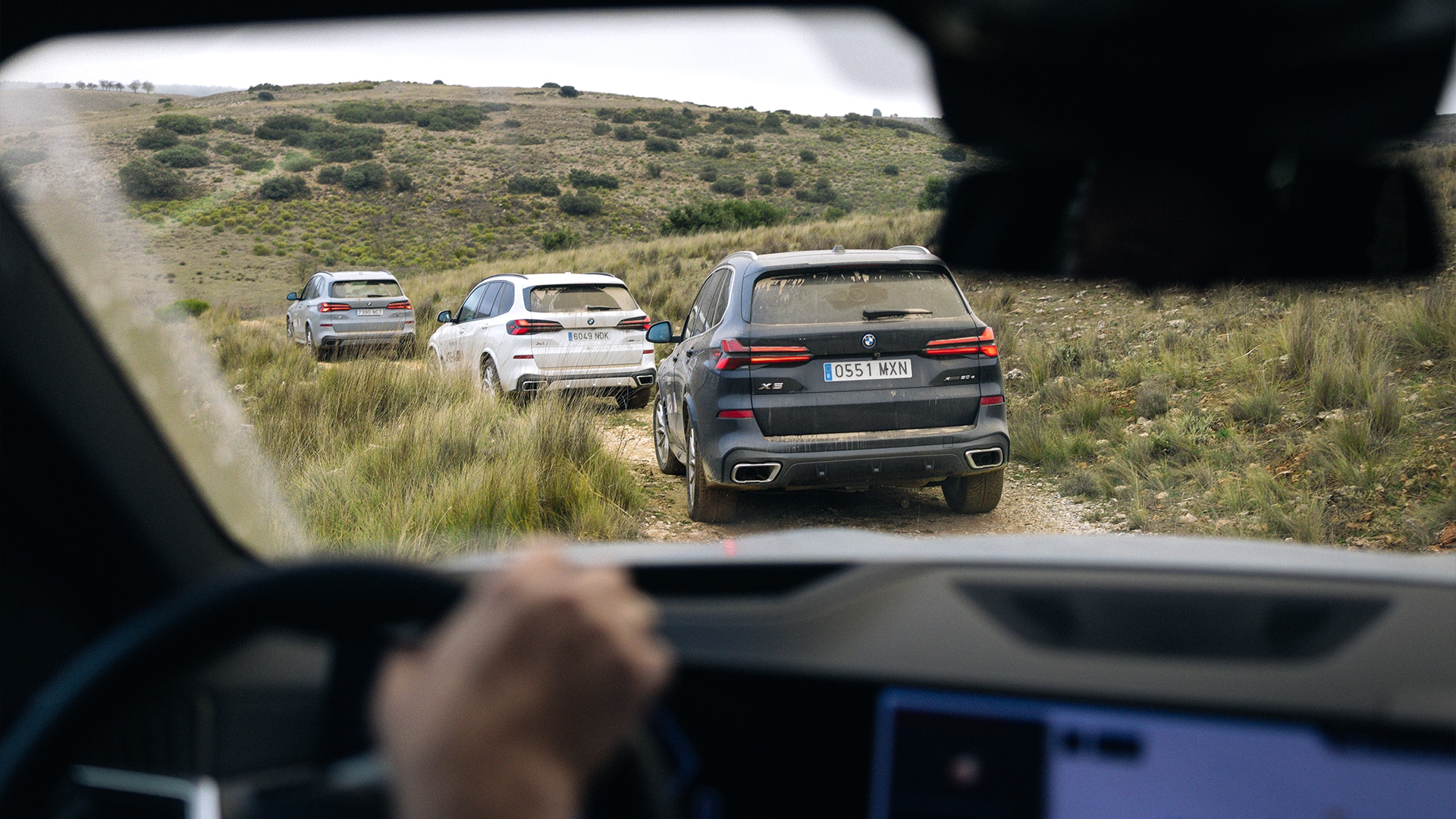 Blick aus dem Inneren eines Fahrzeugs auf drei BMW SUVs, die auf einer Feldstraße in einer hügeligen, grasigen Landschaft fahren. Im Vordergrund ist das Armaturenbrett des Beobachtungsfahrzeugs sichtbar, während drei Fahrzeuge voraus zu sehen sind: ein dunkelgrauer BMW X5 im Vordergrund, ein weißer BMW in der Mitte und ein silberner BMW links. Sanfte Hügel mit spärlicher Vegetation und trockenes Gras erstrecken sich in den Hintergrund.