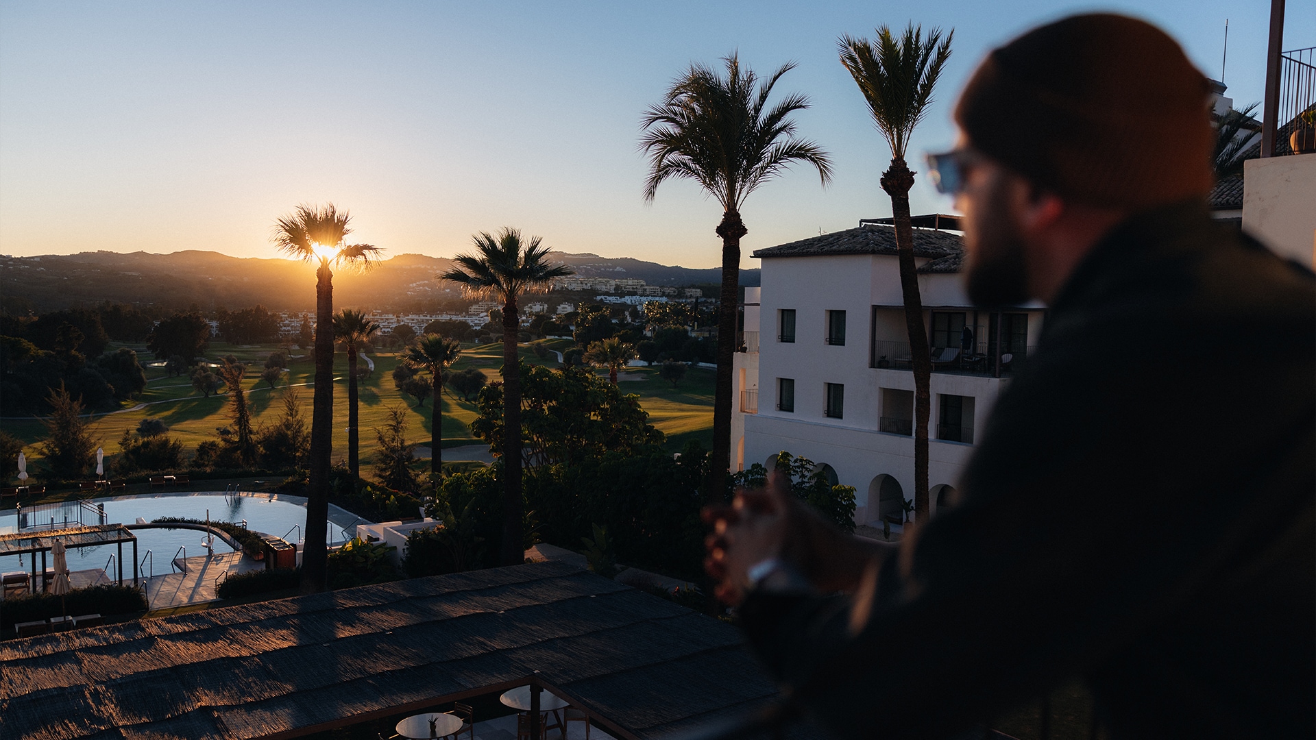 Eine Person in dunkler Kleidung steht auf einer Terrasse mit Blick auf eine Sonnenunterganglandschaft. Palmen rahmen die Aussicht ein, rechts ist ein weißes Gebäude sichtbar. Ein Swimmingpool und ein Golfplatz sind darunter zu sehen, vor Bergen und einem goldenen Sonnenuntergangshimmel.