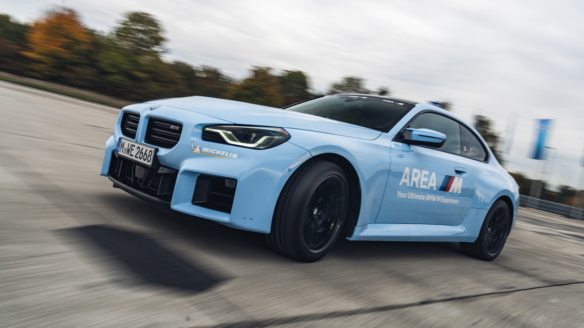 Ein hellblaues BMW Sportauto in Bewegung auf einer Rennstrecke. Das Fahrzeug hat schwarze Räder und trägt die Aufschrift 'AREA M' an der Seite sowie ein Michelin-Logo an der Front. Das Fahrzeug scheint während einer Fahrdemonstration auf einer Betonfläche fotografiert worden zu sein, mit Herbstbäumen im Hintergrund unter einem bewölkten Himmel.