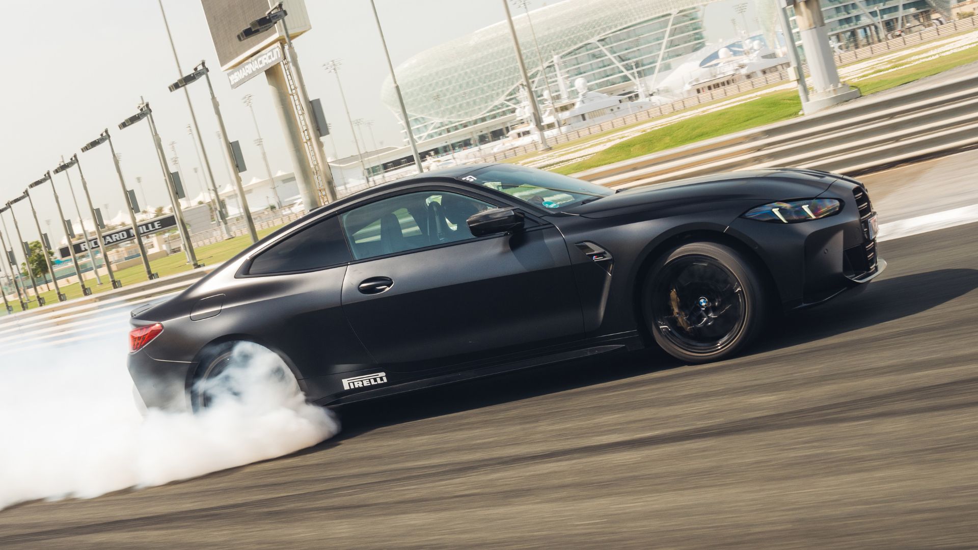 Ein eleganter schwarzer BMW Sportwagen driftet auf einer Rennstrecke und erzeugt weißen Rauch von den Hinterreifen. Das Fahrzeug ist in Bewegung auf einer Strecke mit der markanten Yas Marina Anlage im Hintergrund. Pirelli-Branding ist an der Seite des Autos sichtbar.