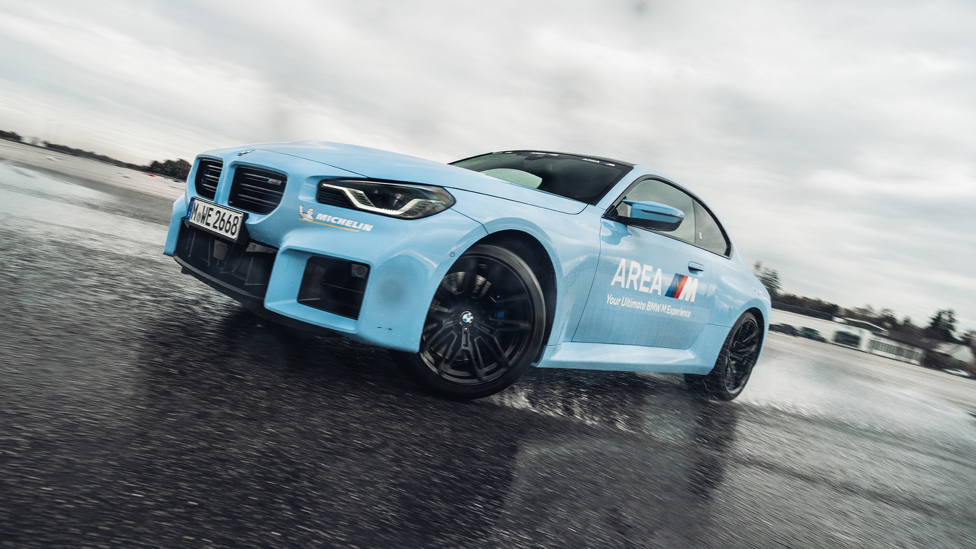 Ein hellblauer BMW Sportwagen mit schwarzen Rädern auf einer nassen Fahrstrecke. Das Fahrzeug trägt die Aufschrift 'AREA M' an der Seite und ein Michelin-Logo an der Front. Es scheint in Bewegung auf nassem Asphalt unter bewölktem Himmel fotografiert zu sein.