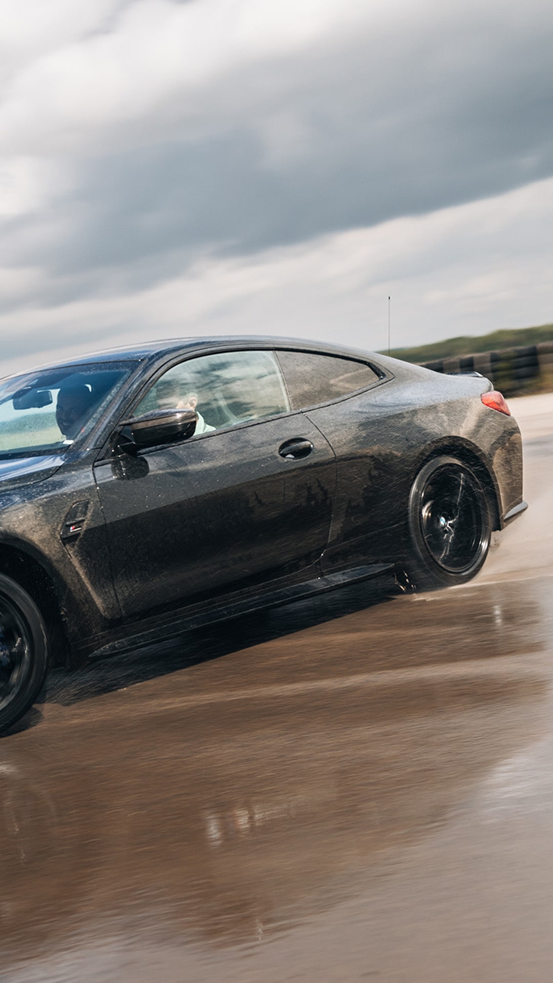 Ein elegantes schwarzes Sportcoupé, das auf einer nassen Straße unter bewölktem Himmel fährt. Das Fahrzeug hat schwarze Leichtmetallfelgen und scheint in Bewegung zu sein, wobei Wasser unter den Reifen spritzt.