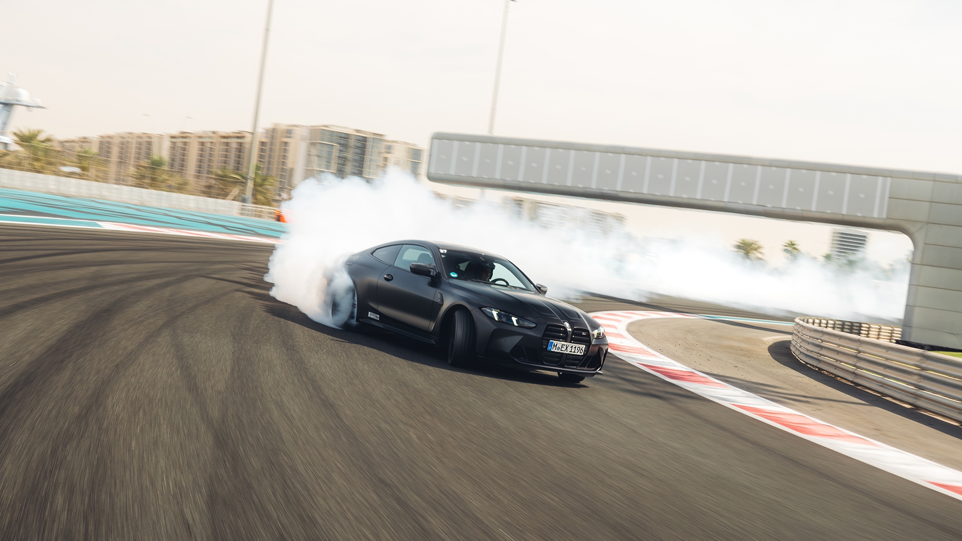Ein schwarzer BMW Sportwagen driftet auf einer Rennstrecke und erzeugt weißen Rauch von den durchdrehenden Reifen. Das Fahrzeug navigiert durch eine Kurve mit rot-weißen Streckenmarkierungen. Im Hintergrund sind Gebäude und ein klarer Himmel zu sehen.