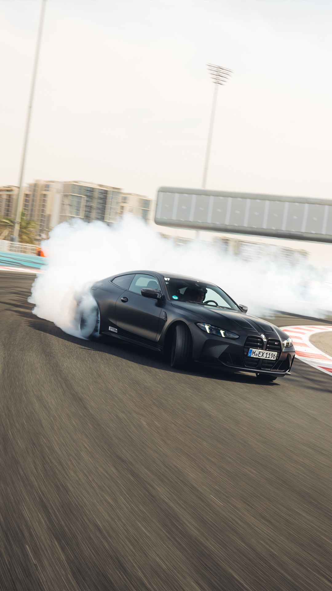 Ein mattschwarzer BMW Sportwagen driftet auf einer Rennstrecke und erzeugt eine Wolke aus weißem Reifenrauch. Das Fahrzeug fährt auf einer Asphaltbahn mit Stadionlichtern und Gebäuden im Hintergrund unter einem hellen Himmel.