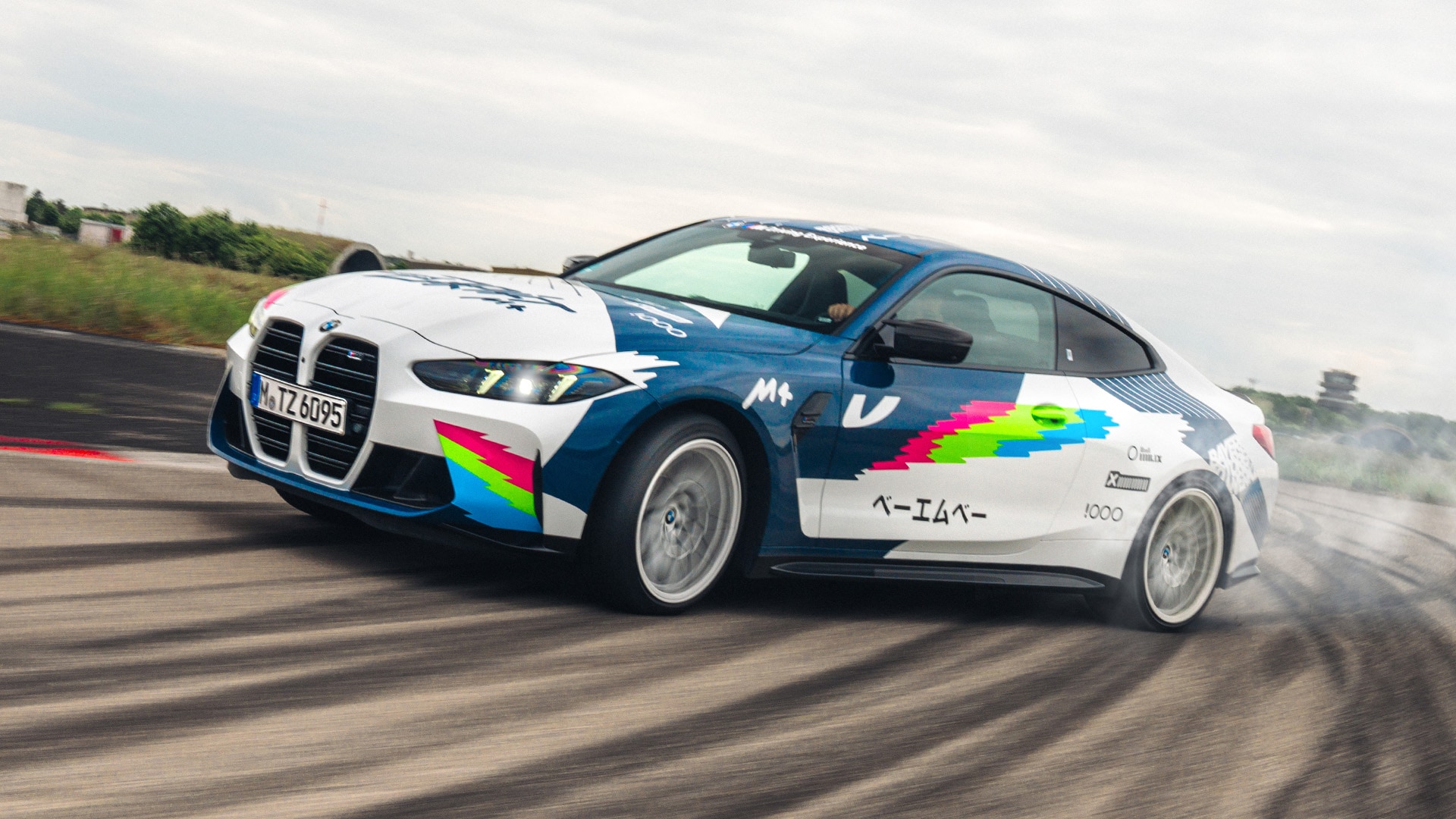 Ein BMW Sportwagen mit einer markanten blau-weißen Farbgebung und bunten Grafiken beim Driften auf einer Rennstrecke. Das Fahrzeug hat leuchtende rosa, grüne und blaue Akzente an der Seite und japanische Schriftzeichen aufgedruckt. Reifenspuren sind auf dem Asphalt sichtbar, während das Auto unter bewölktem Himmel das Drift-Manöver ausführt.