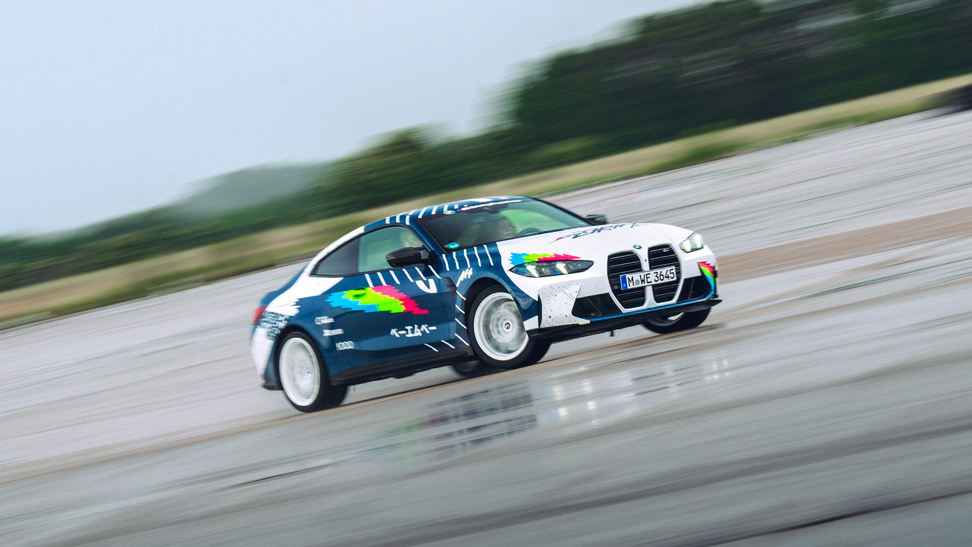Ein BMW Sportwagen mit einer markanten blauen, weißen und bunten Grafik-Folierung fährt auf einer breiten, flachen Betonfläche. Das Fahrzeug scheint in Bewegung zu sein, mit drehenden Rädern, was ein Gefühl von Geschwindigkeit vor dem verschwommenen Hintergrund aus Bäumen und Himmel erzeugt.