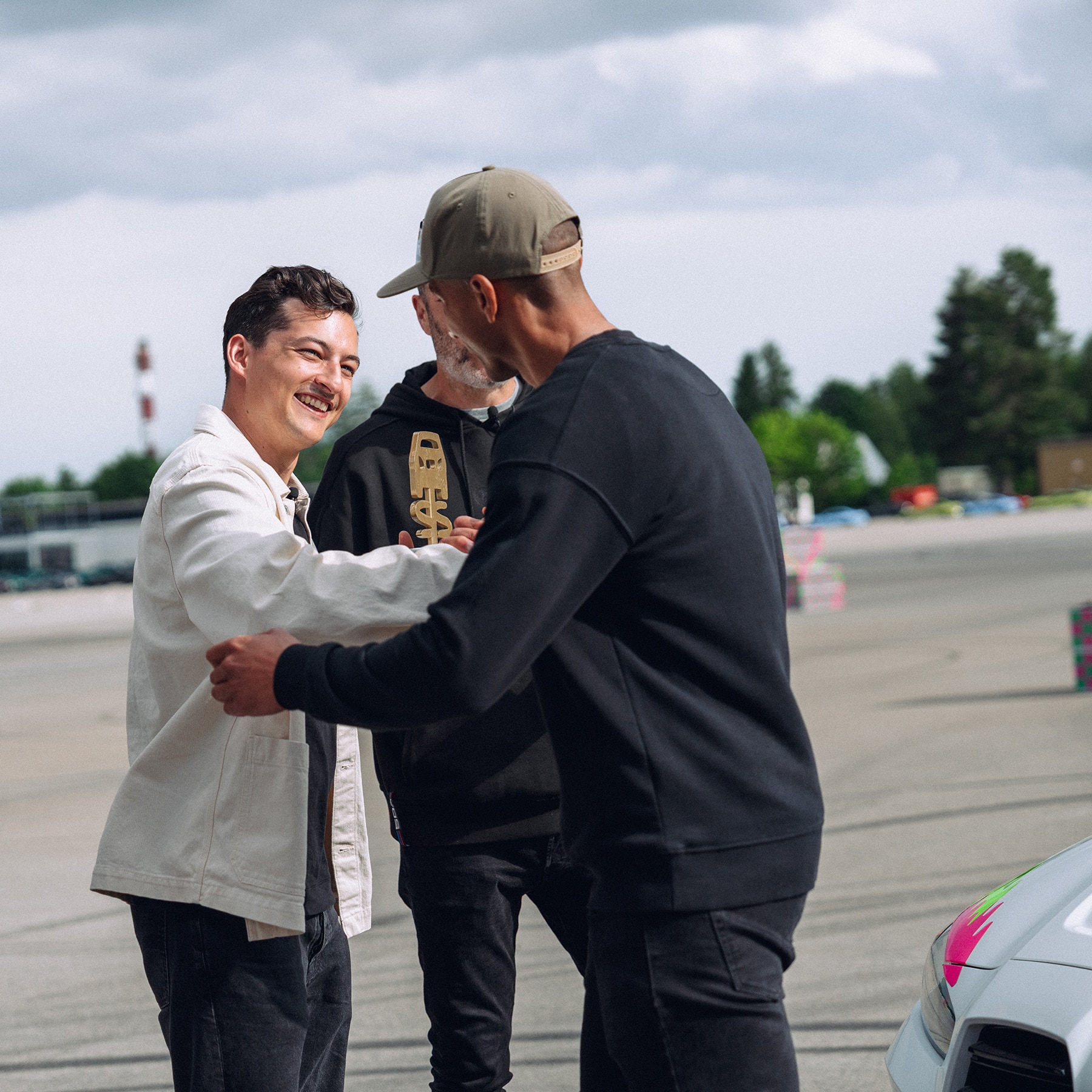 Drei Personen begrüßen sich im Freien auf einem Parkplatz oder einer offenen Fläche. Eine Person trägt eine weiße Jacke, während eine andere dunkle Kleidung mit einer Mütze trägt. Im Hintergrund sind Bäume und ein bewölkter Himmel zu sehen, mit etwas, das wie ein Leuchtturm oder ein Turm in der Ferne aussieht.