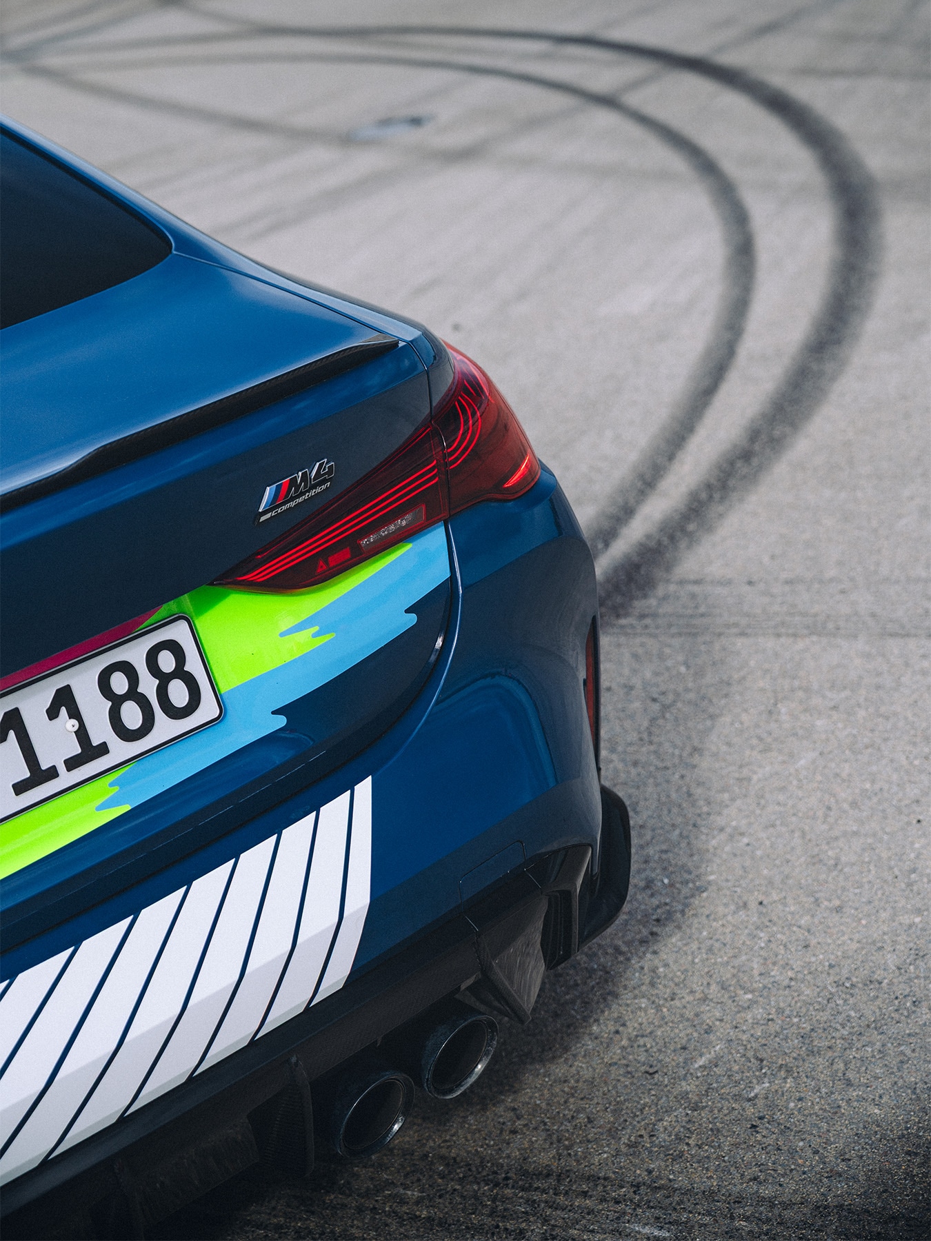Rückansicht eines blauen BMW M4 Sportwagens mit farbigen Akzenten und einem teilweise sichtbaren Kennzeichen. Im Hintergrund sind dunkle Reifenspuren auf der Betonoberfläche zu sehen, die auf kürzliches Driften oder sportliches Fahren hindeuten.