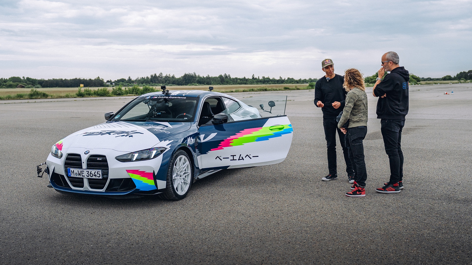 Ein weißer BMW Sportwagen mit bunten rosa, grünen und blauen Grafiken und montierten Kameras steht auf einem offenen Asphalt. Drei Personen stehen neben dem Fahrzeug und betrachten es, wobei eine geöffnete Tür das Innere zeigt. Die Szene spielt auf einer Teststrecke mit offener Landschaft im Hintergrund unter bewölktem Himmel.