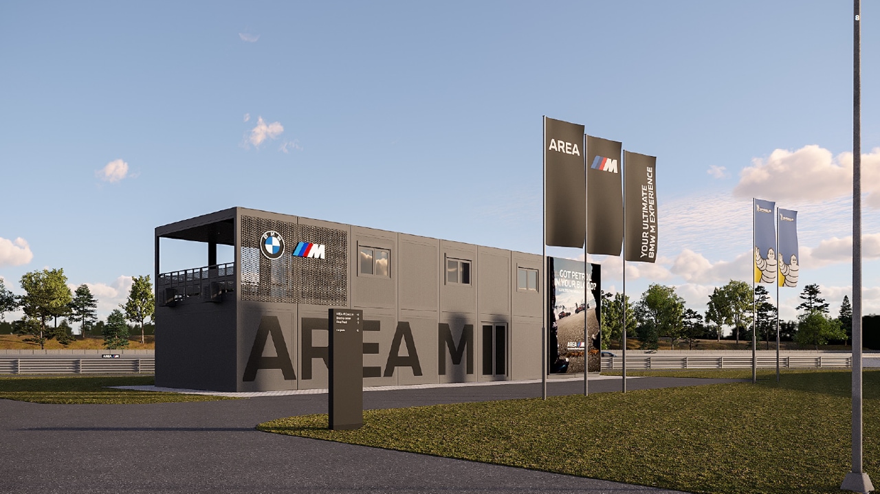 Eine moderne BMW M-Einrichtung namens 'AREA M' mit einem grauen zweistöckigen Gebäude, das das BMW-Logo trägt. Mehrere hohe Banner zeigen 'AREA M' und 'YOUR ULTIMATE BMW EXPERIENCE'. Das Gebäude befindet sich in der Nähe einer Rennstrecke mit Bäumen im Hintergrund unter einem klaren blauen Himmel.