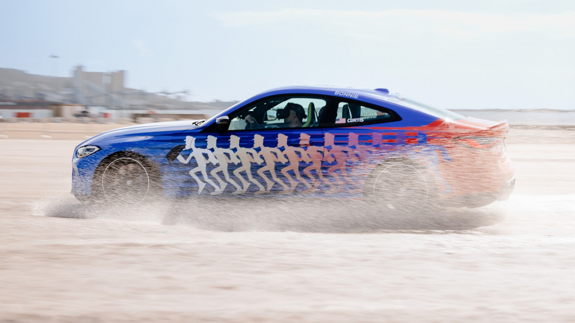 Ein blaues Sportauto mit einem Farbverlauf, der am Heck in Rot übergeht, rast über eine sandige Oberfläche und wirbelt Staub auf. Das Fahrzeug ist mit einem dekorativen Muster aus laufenden Figuren an der Seite verziert. Das Fahrzeug ist in Bewegung auf dem, was wie eine Wüste oder ein Strandbereich aussieht, mit einem klaren blauen Himmel im Hintergrund.