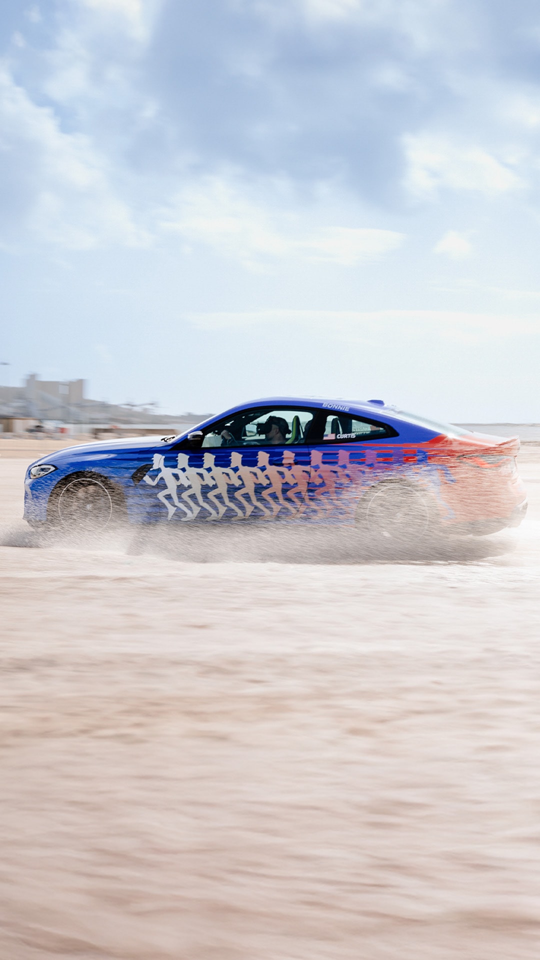 Ein blauer Sportwagen mit einem markanten Grafik-Design, das laufende Figuren-Silhouetten zeigt, fährt über einen Sandstrand und wirbelt Sprühwasser auf. Das Fahrzeug scheint sich auf nassem Sand unter einem teilweise bewölkten Himmel zu bewegen.