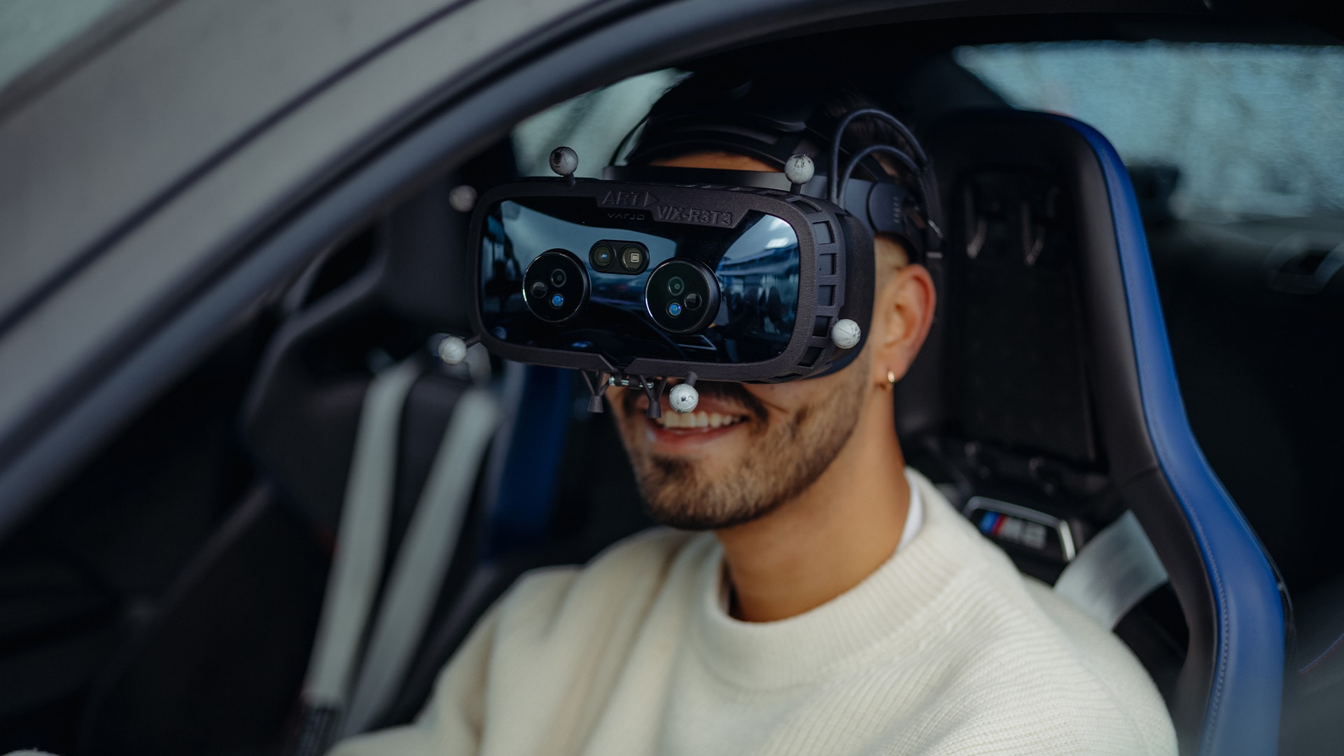 Eine Person sitzt in einem Fahrzeug mit blauen Innensitzen und trägt ein Virtual-Reality-Headset. Die Person trägt einen weißen Pullover und scheint zu lächeln, während sie das VR-Gerät benutzt, das mehrere sichtbare Sensoren oder Kameras an der Vorderseite hat.