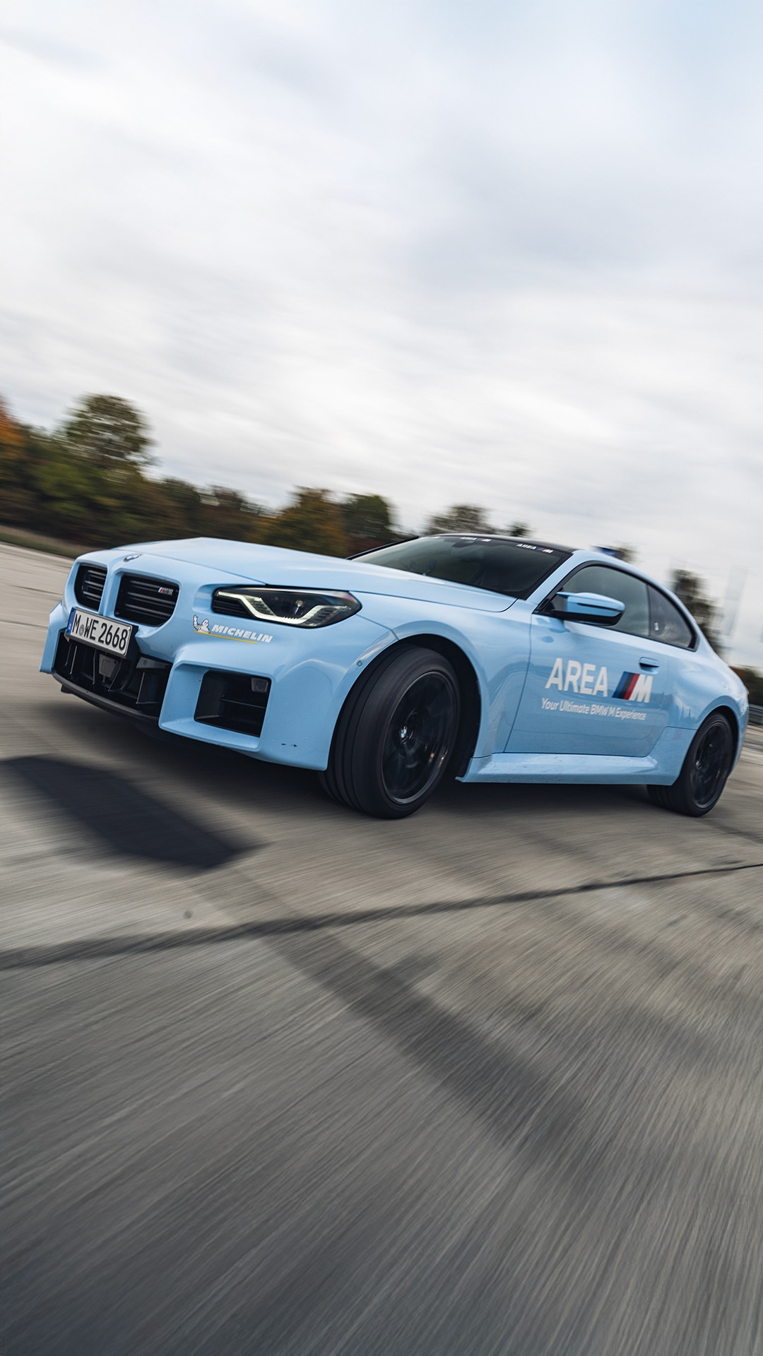 A light blue BMW sports car with 'AREA M' branding and Michelin sponsorship in motion on a track. The vehicle has black wheels and a modified front bumper, captured with motion blur against an overcast sky.
