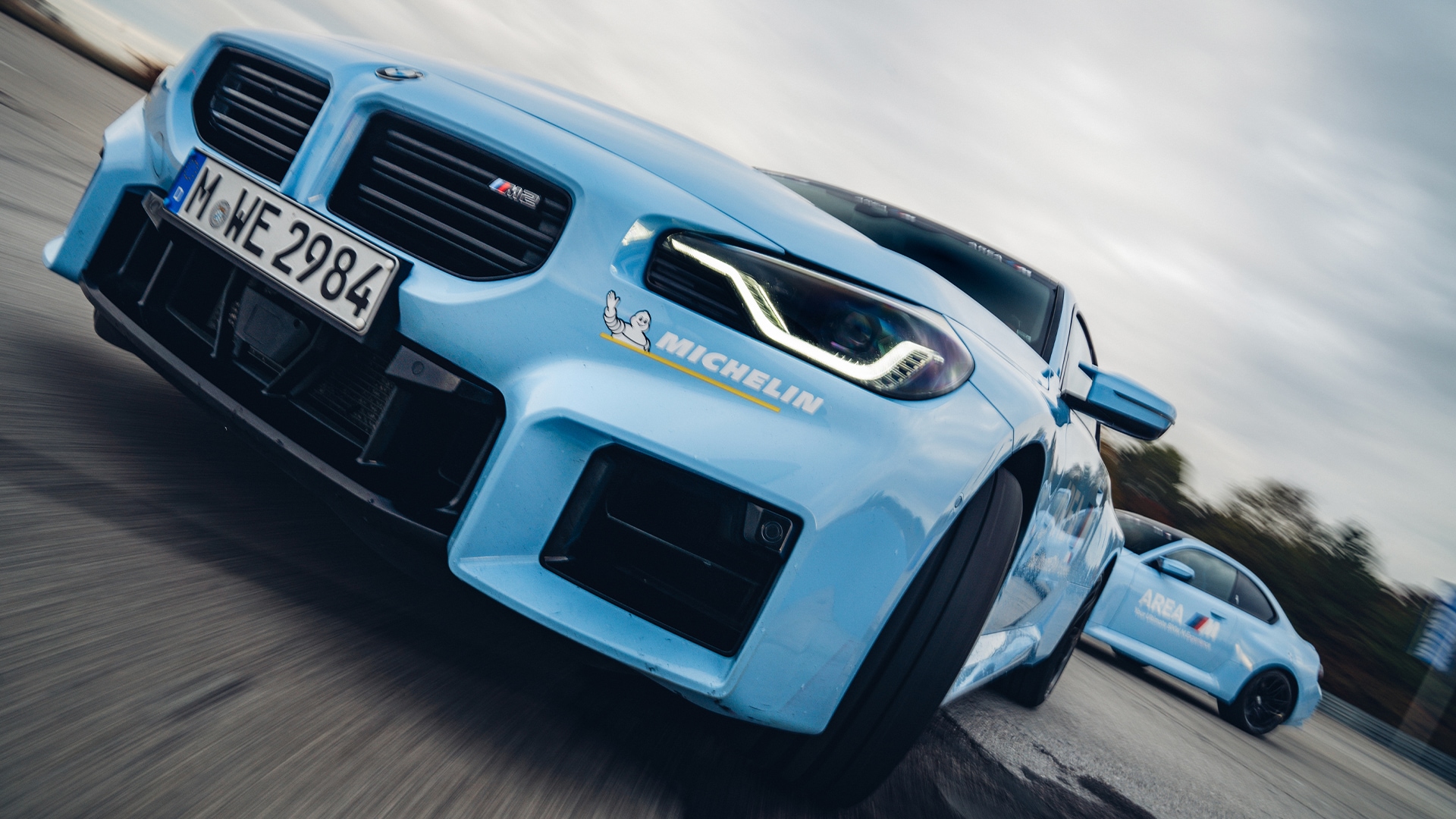 Ein hellblauer BMW Sportwagen mit einem Michelin-Logo an der Seite wird in Bewegung auf einer Straße aufgenommen. Das Fahrzeug hat markante LED-Scheinwerfer und ein deutsches Kennzeichen. Ein zweites blaues Fahrzeug ist im Hintergrund zu sehen.